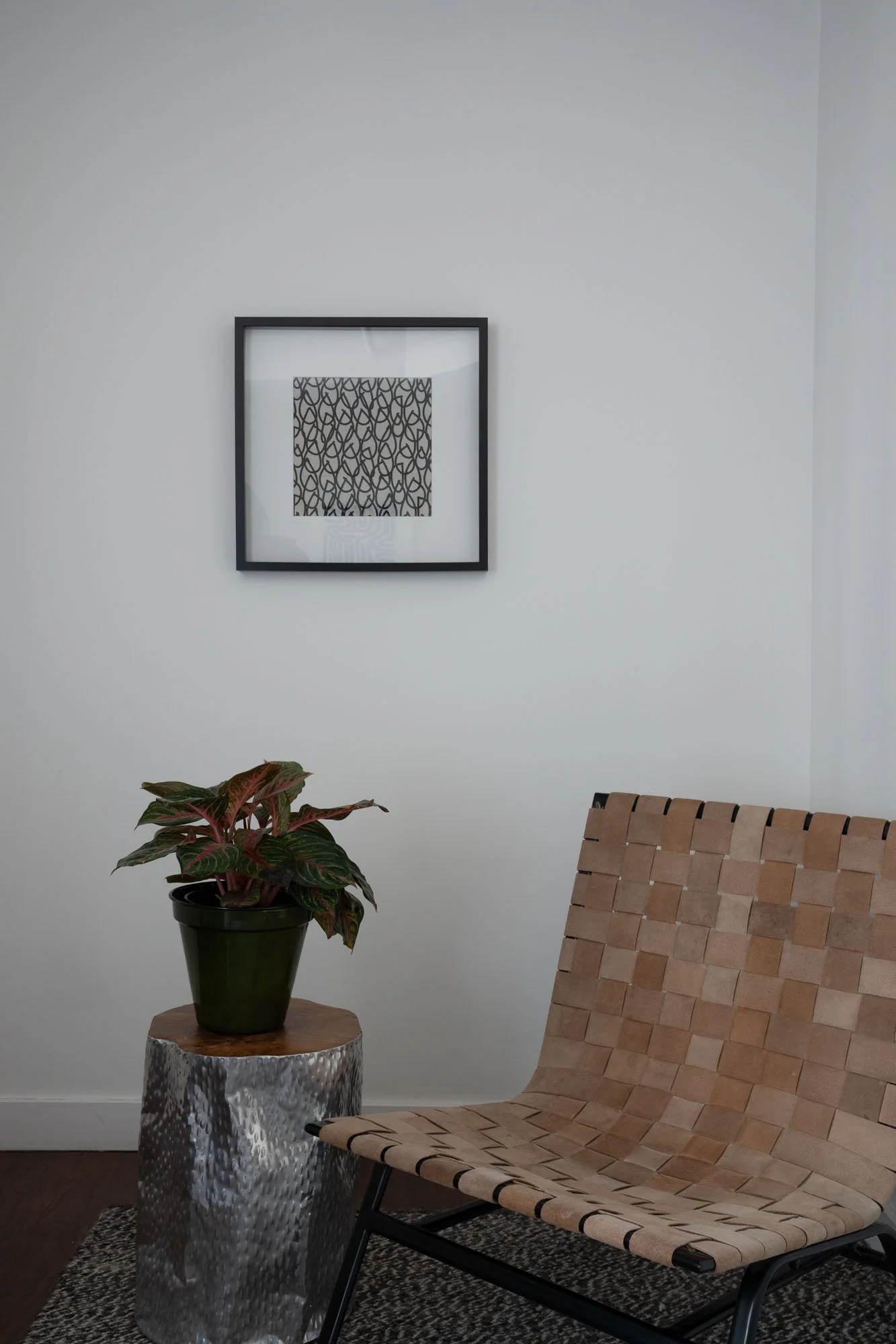 A minimalistic interior space with a framed black-and-white patterned artwork on a white wall, a potted plant on a silver textured side table, and a woven beige lounge chair on a dark rug.