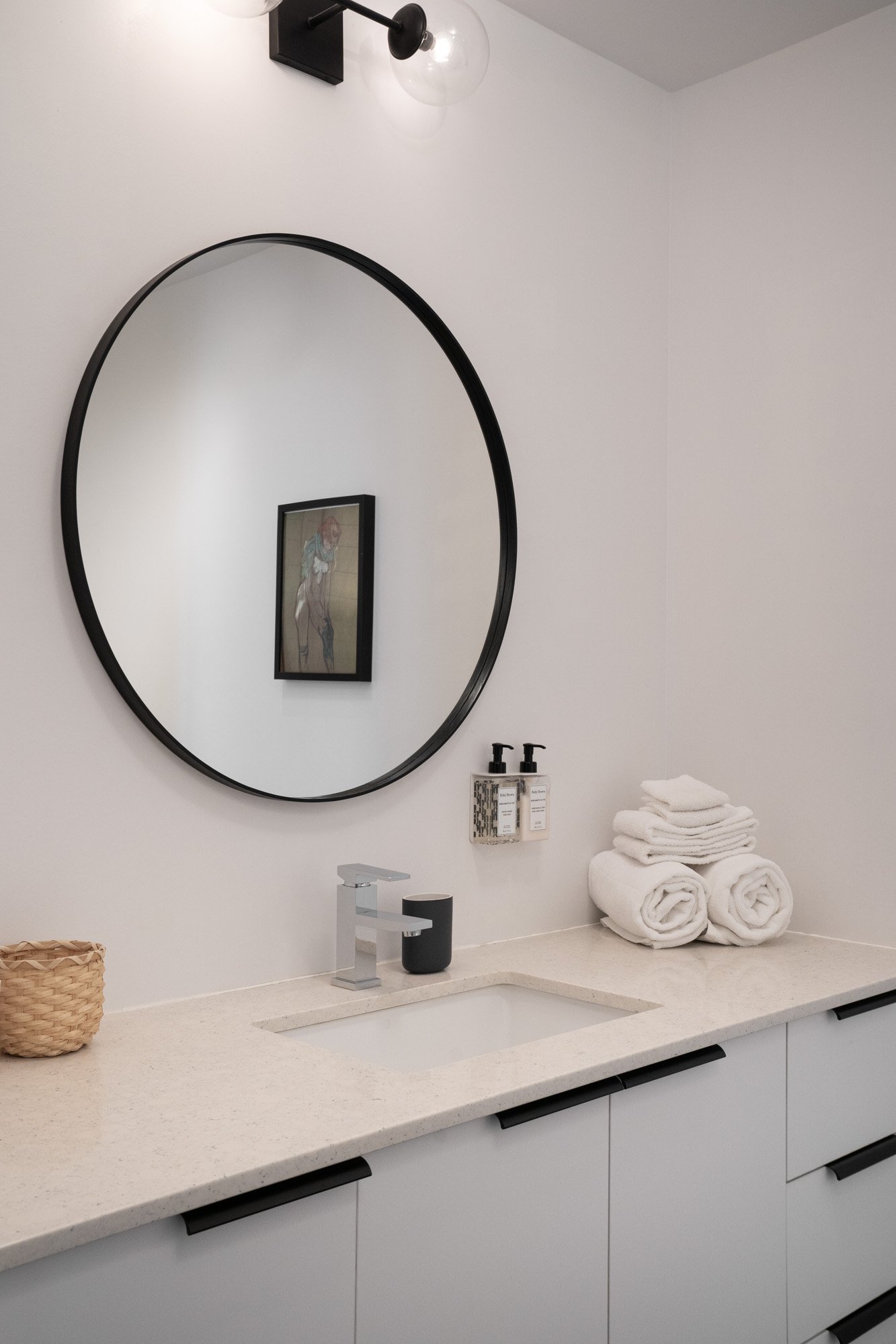 A bathroom vanity with a white countertop, a modern faucet, basket, soap dispenser, neatly folded towels, a wall-mounted soap holder, a large round mirror with a black frame, and a wall-mounted light fixture.