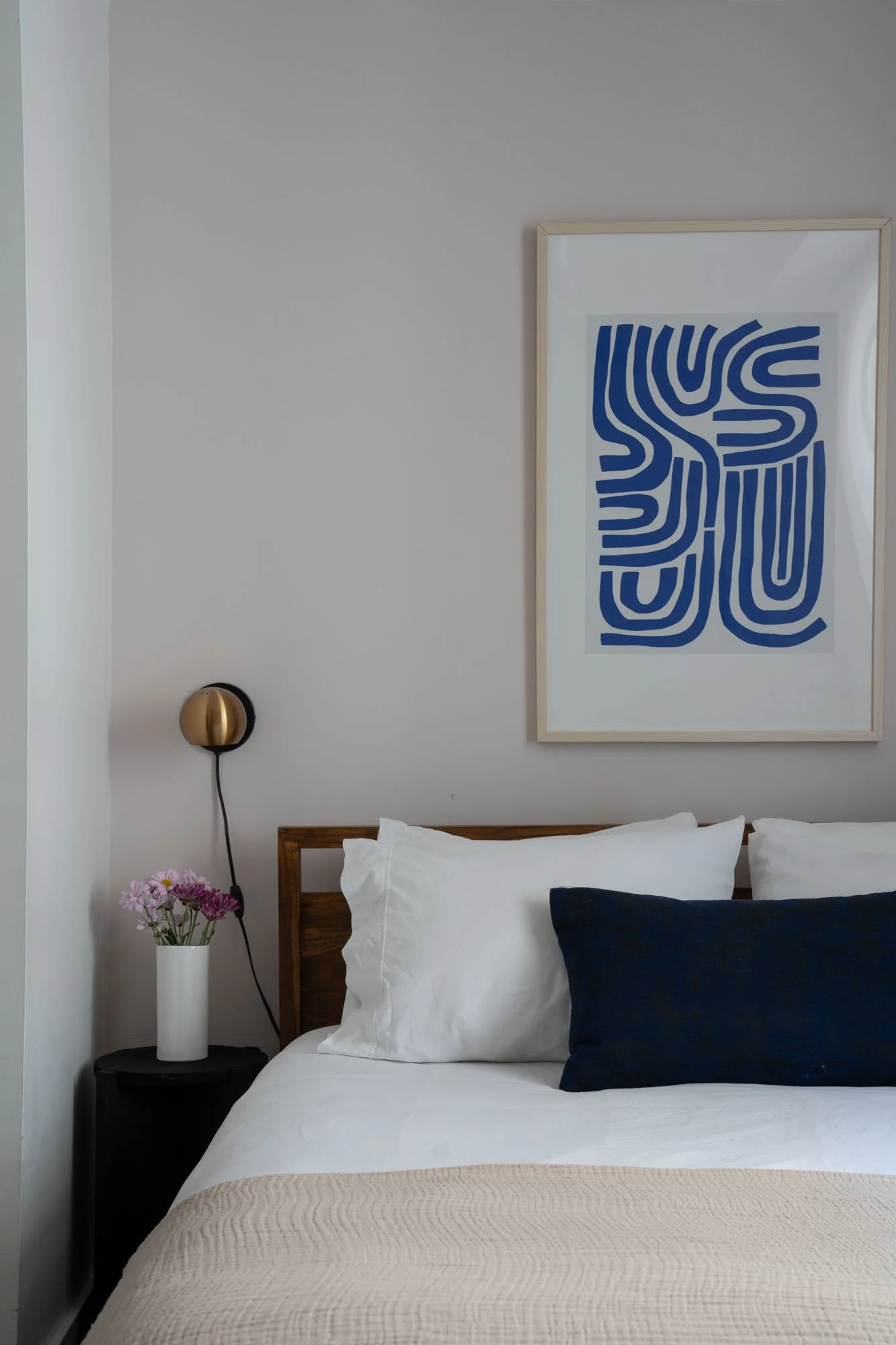 A neatly made bed with white sheets, white pillows, and a navy blue pillow in a bedroom with modern decor. There is a black bedside table with a white vase of pink and purple flowers, a gold and black wall-mounted light, and a framed blue abstract ar