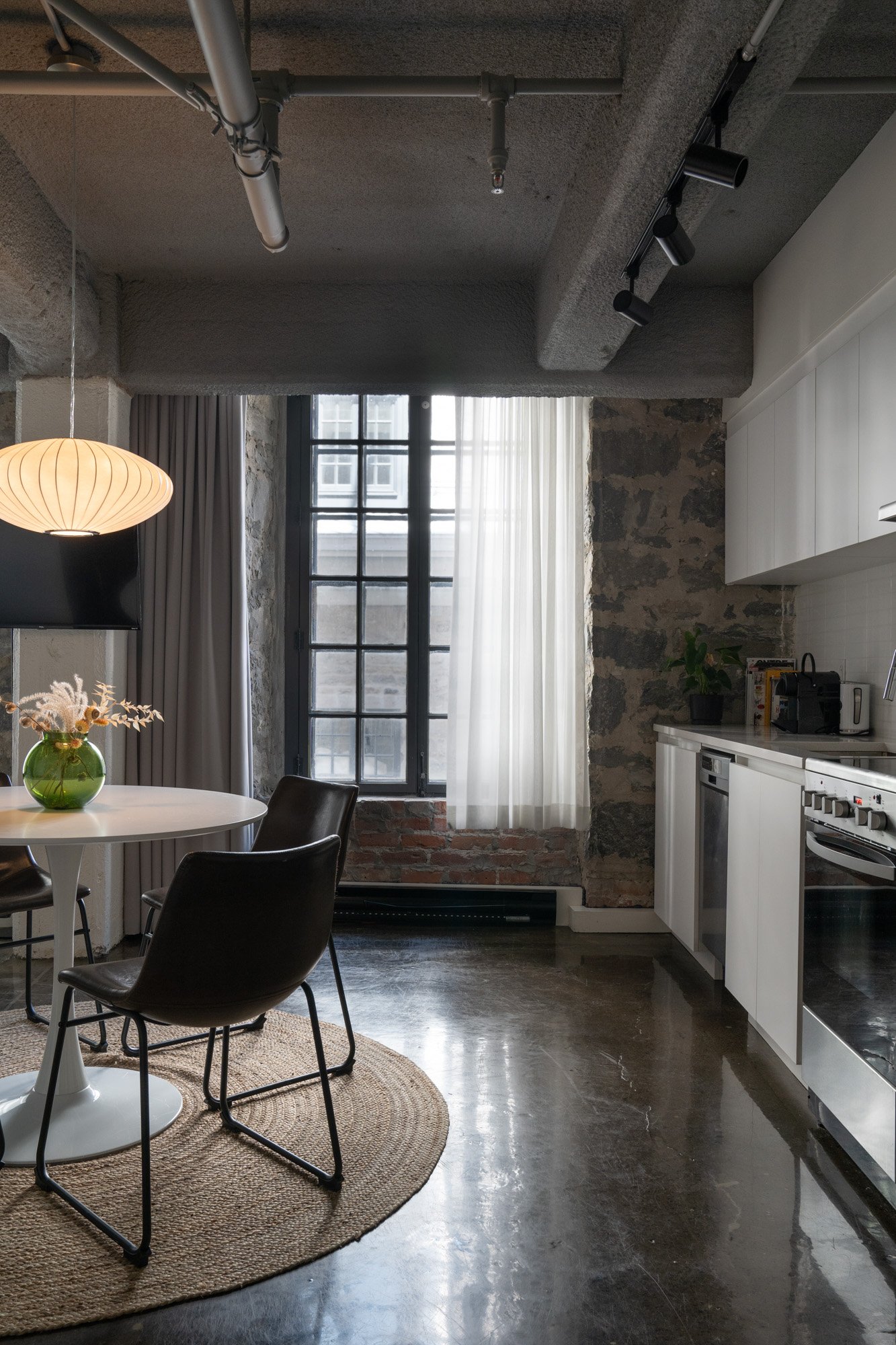 Modern kitchen and dining area with concrete floor, brick wall, large window with sheer curtains, black chairs around a white dining table with a green vase of flowers, white kitchen cabinets, and stainless steel appliances.