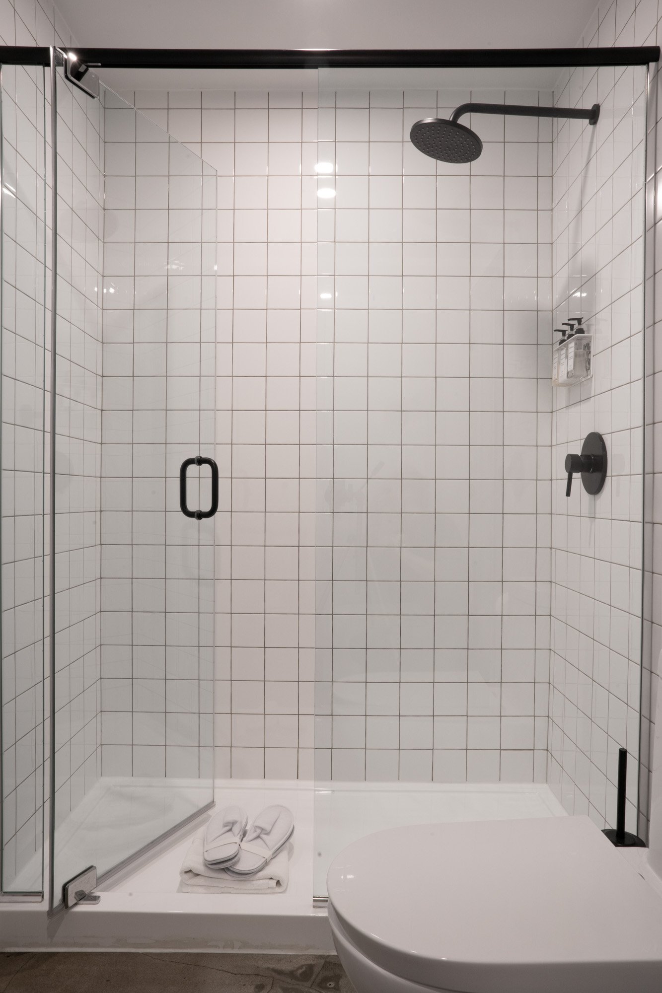 A modern bathroom with a glass-enclosed shower, white tiled walls, a mounted rain showerhead, soap dispensers, towel, and slippers on the shower floor.