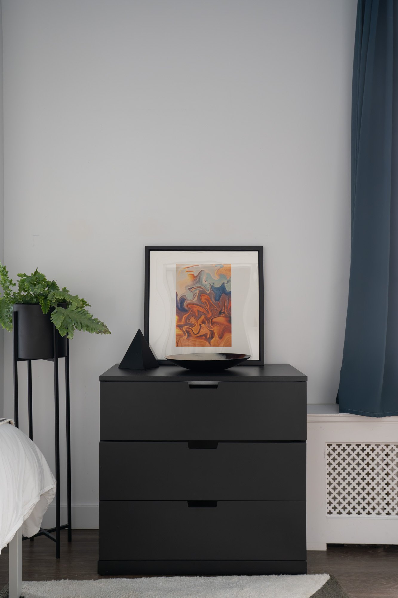 Black dresser with a framed abstract art piece, a black bowl, and a black geometric sculpture on top. To the left, a tall plant stand with a green plant. Part of a bed and a white rug are visible in a room with white walls and blue curtains.