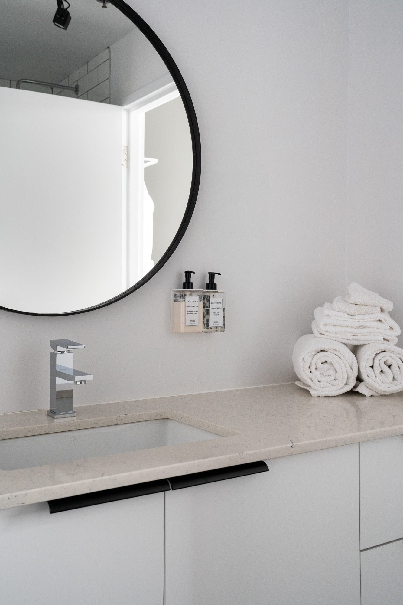 Bathroom vanity with a round mirror, stacked rolled white towels, soap dispensers, and a modern faucet.
