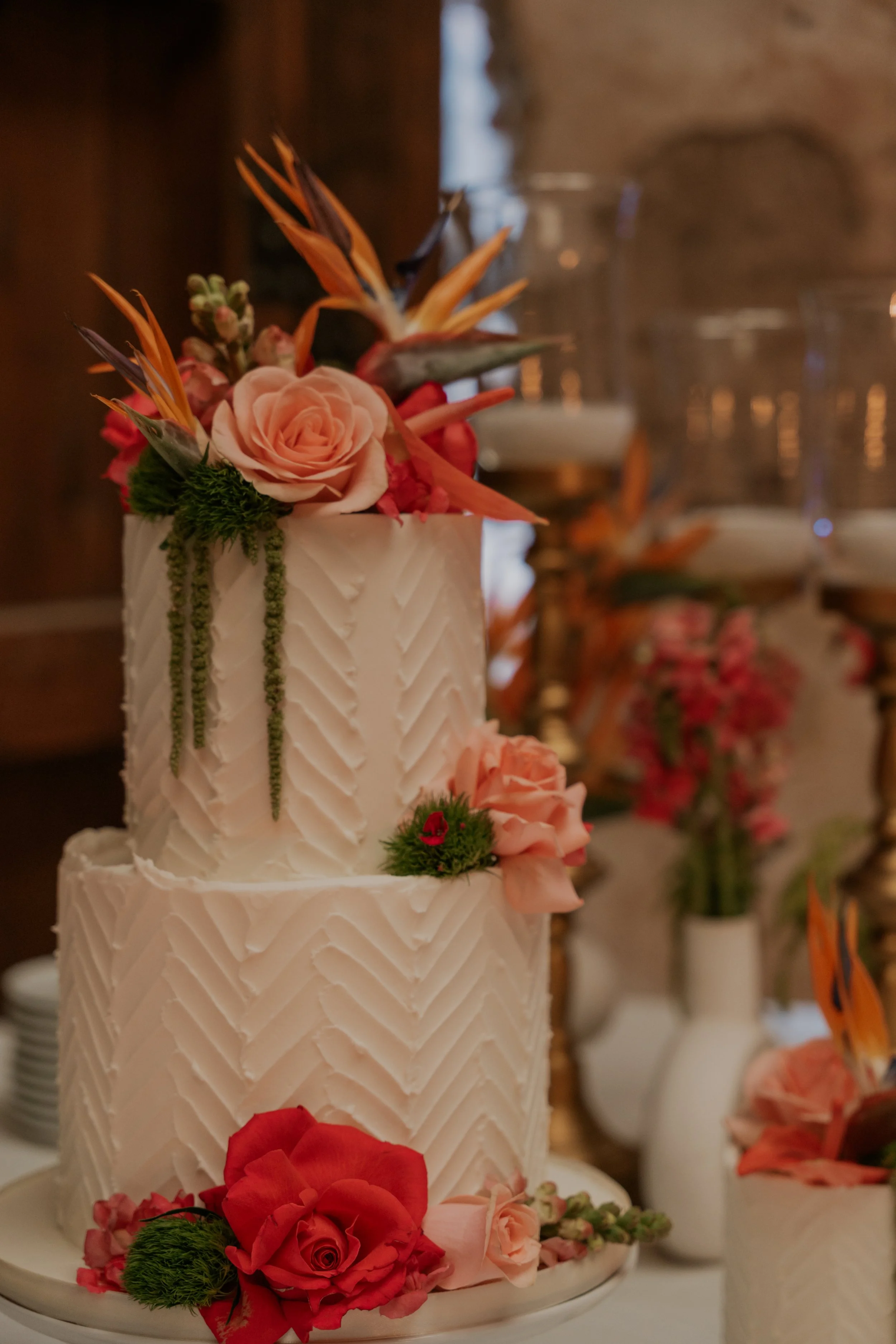 Two-tiered wedding cake decorated with pink and red roses, green foliage, and orange bird of paradise flowers, set on a white platter.