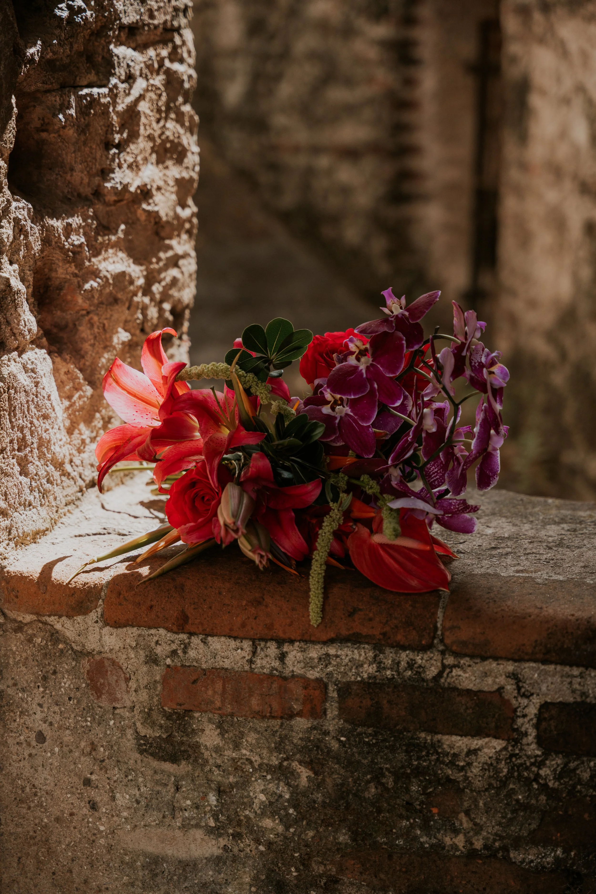 Bouquet of red, purple, and pink flowers resting on a weathered brick ledge against an old brick wall.