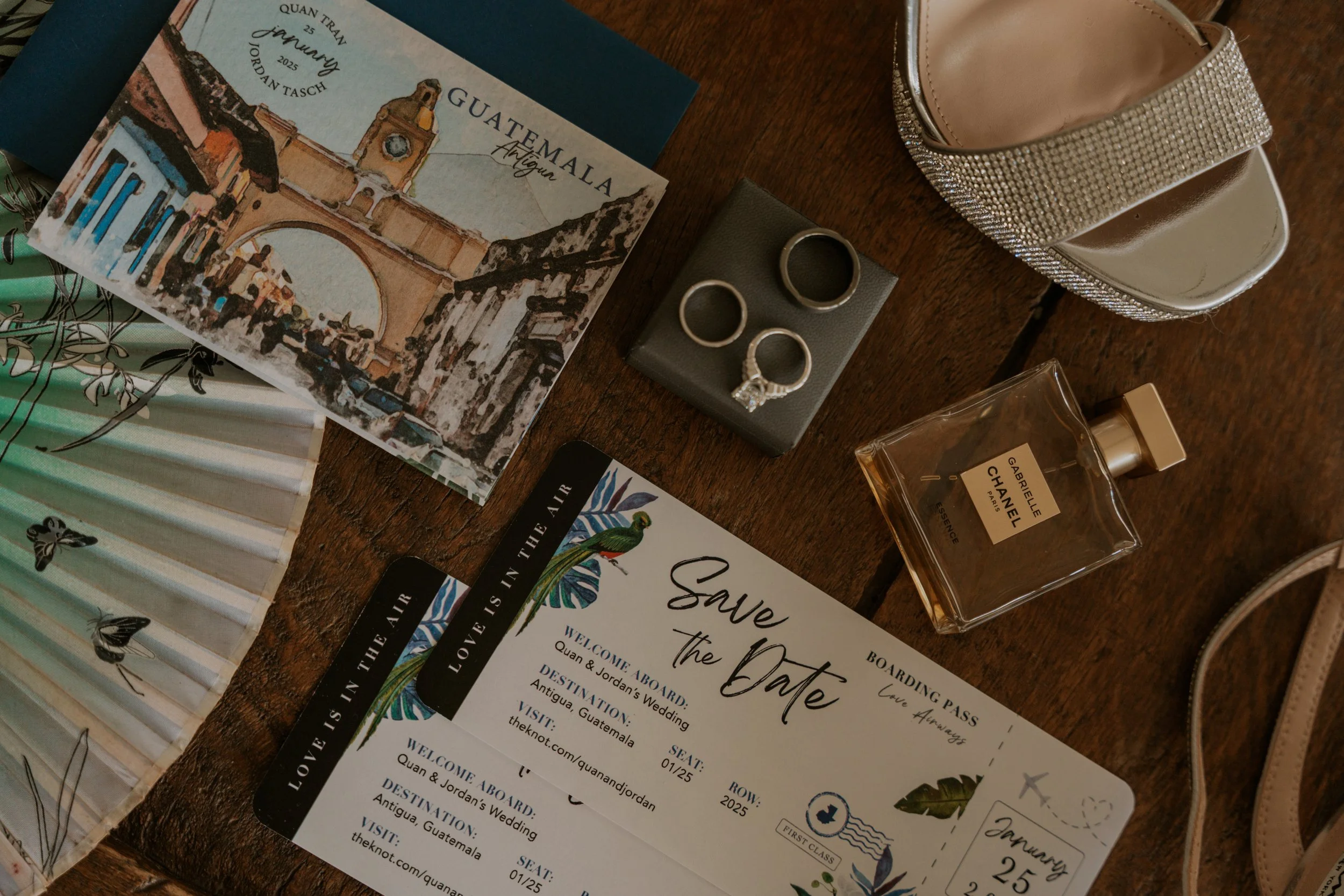 Guatemalan travel brochure, wedding rings in a box, Chanel perfume bottle, white and rhinestone embellished shoes, save-the-date card, and a fan with butterfly print on a wooden table.