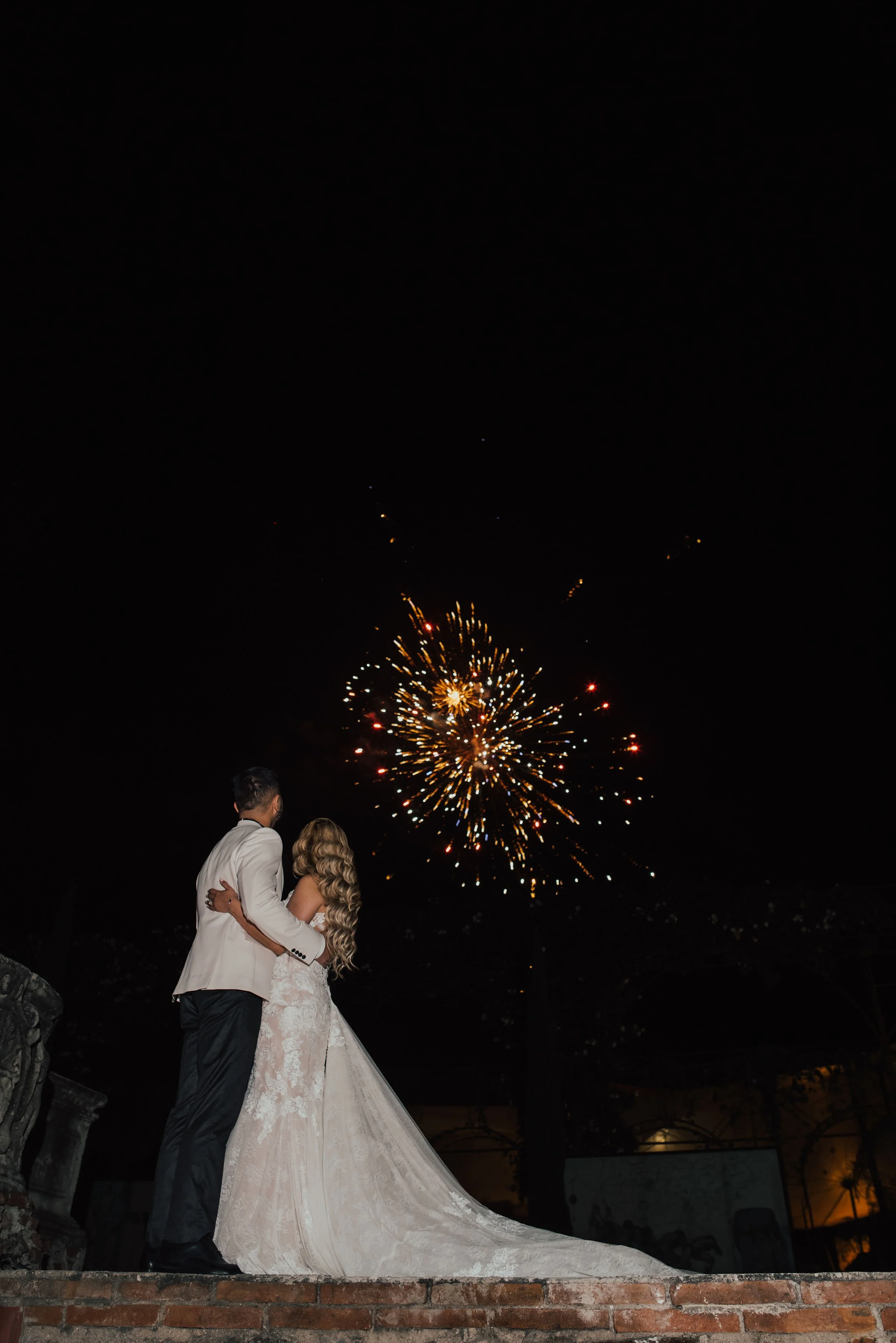 Bride and groom in wedding attire embracing outdoors at night, watching fireworks in the sky.