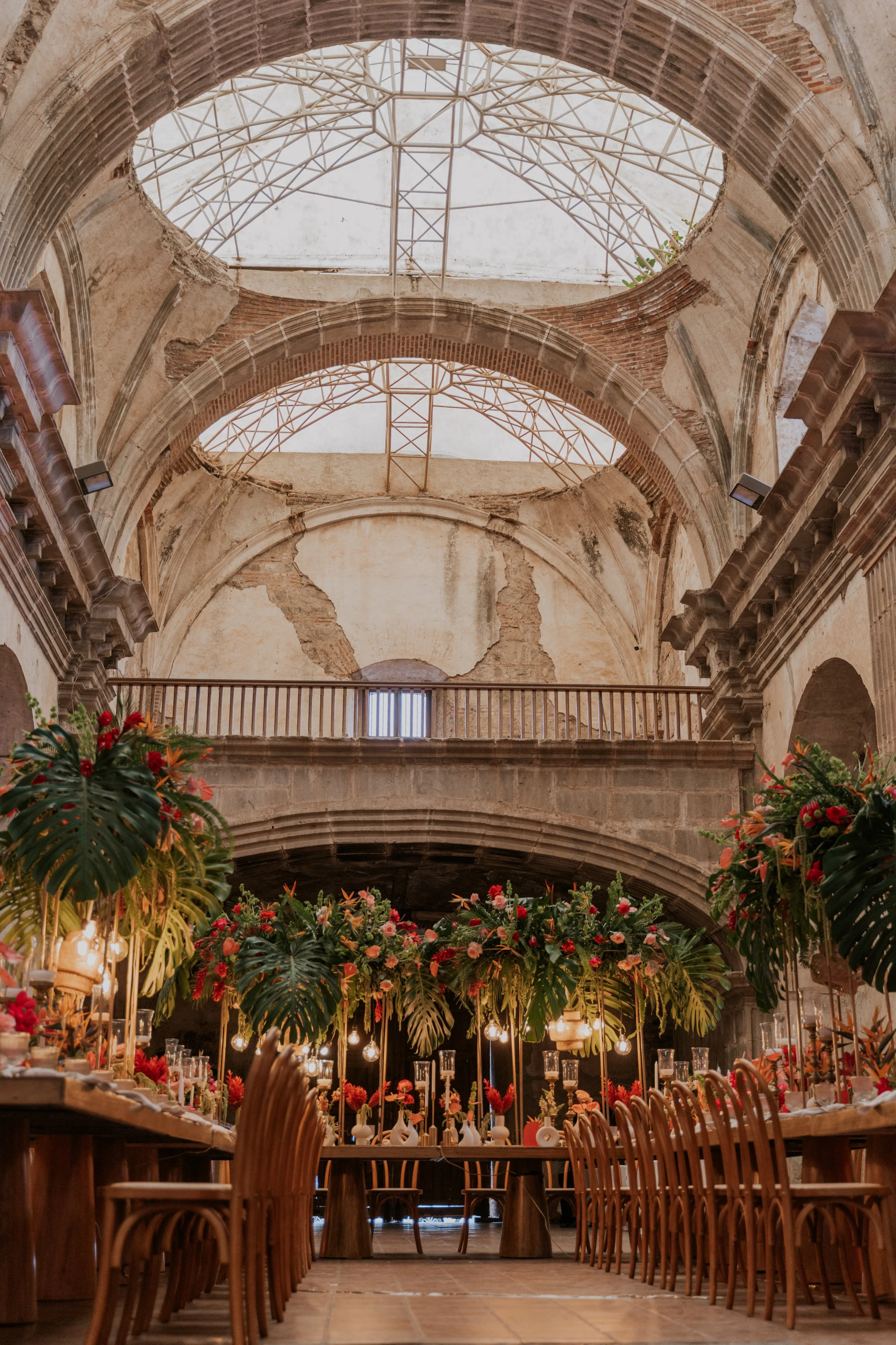 Inside a historic church with exposed brick walls and a high arched ceiling, decorated for an event with large tropical green leaves and vibrant flowers, set tables, and chairs.