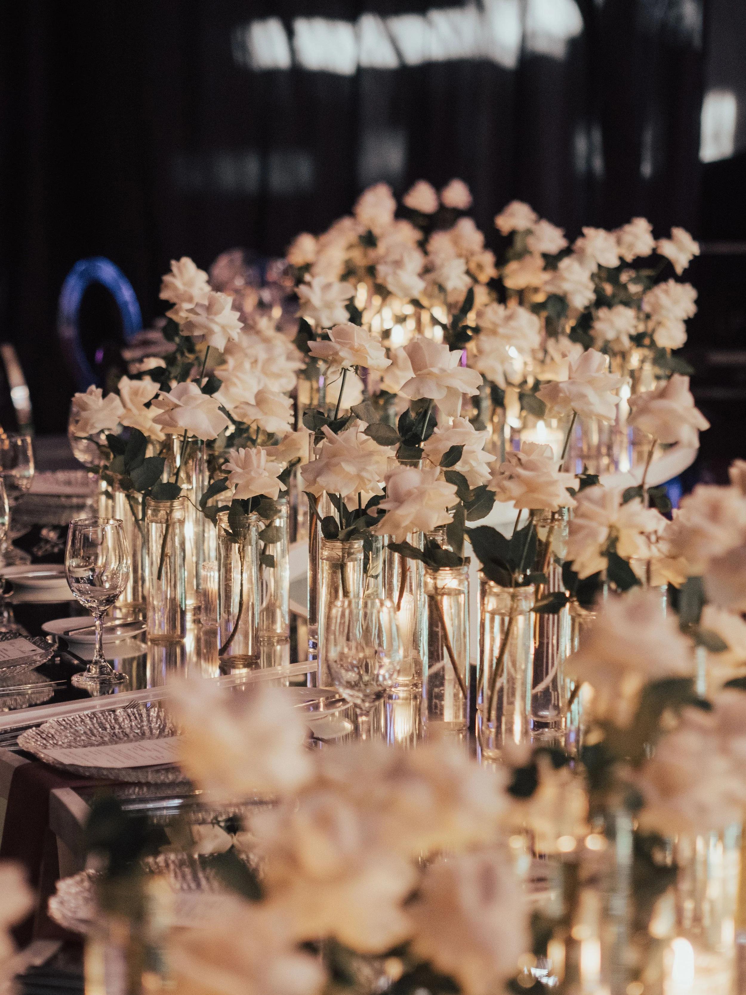 Elegant dining table with white roses in glass vases, candlelight, and glassware for a formal event.