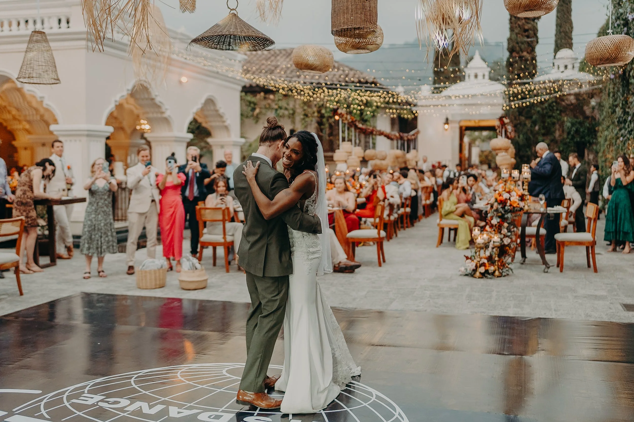 A bride and groom are dancing at their wedding reception with guests watching and taking photos in the background, decorated with string lights and floral arrangements.