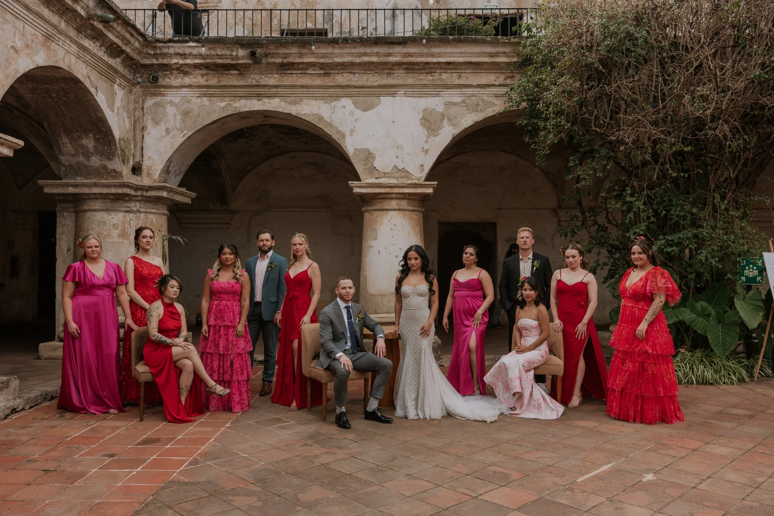 Group of people at a wedding in an outdoor courtyard, with some seated and others standing, dressed in formal attire, with a rustic stone wall and greenery in the background.