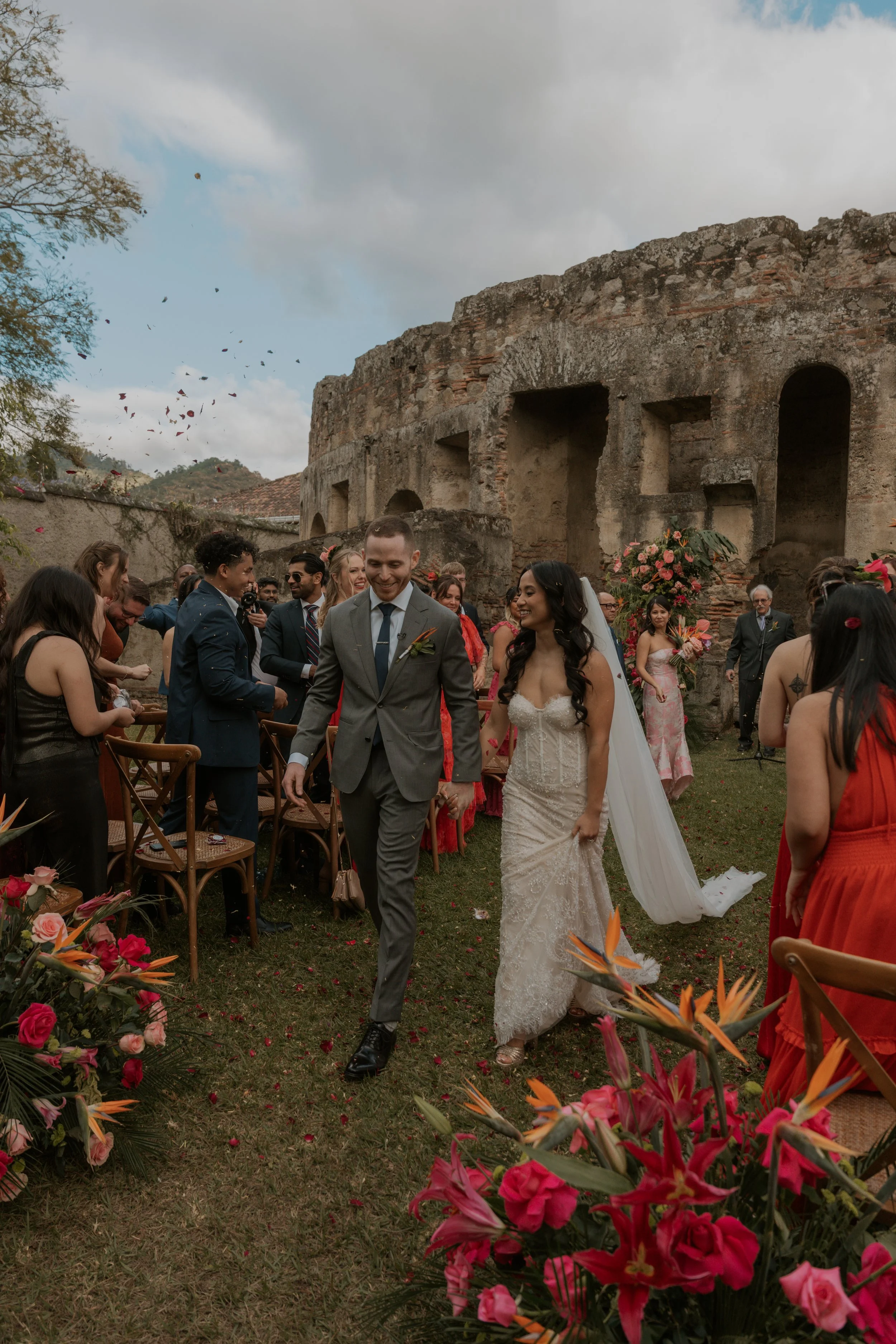 A bride and groom walking hand in hand through a wedding reception outdoors. The setting includes an ancient stone structure in the background, colorful flowers in the foreground, and guests dressed in formal attire celebrating around them.
