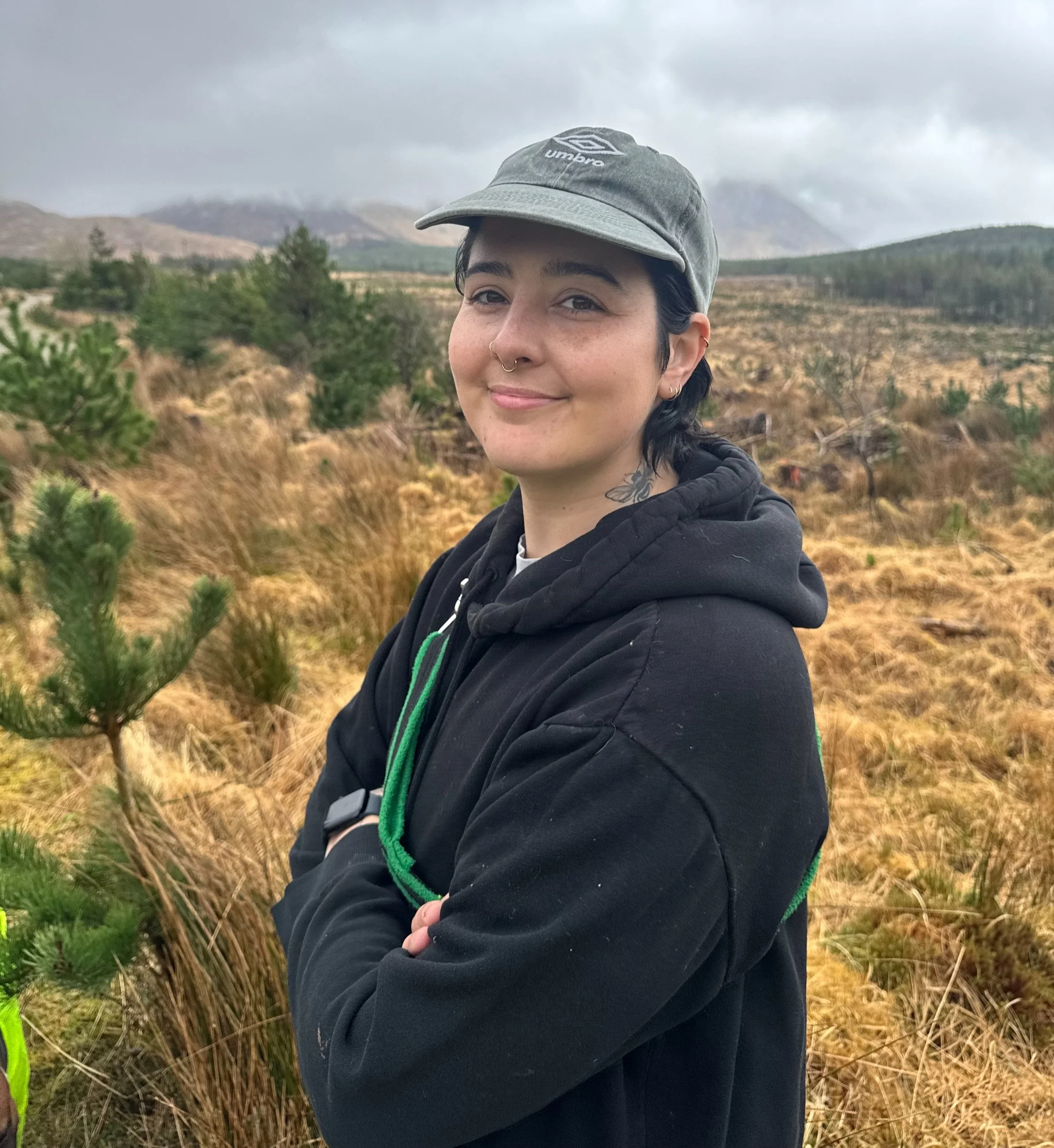 A young person with dark hair, wearing a gray cap and black hoodie, smiling with folded arms outdoors in a grassy, mountainous area with cloudy skies.