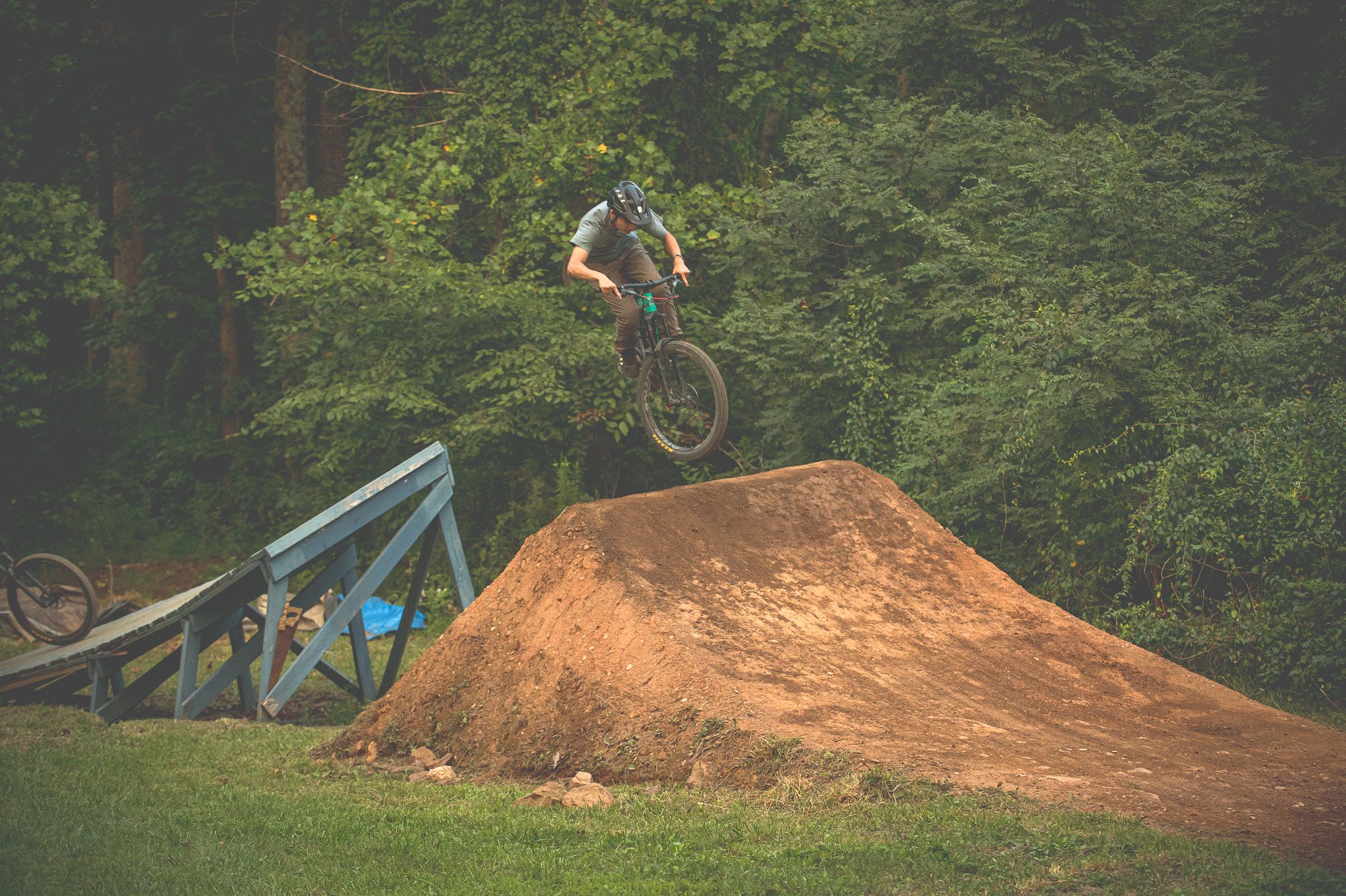 Mountain biker performing a jump over a dirt ramp in a forested area.