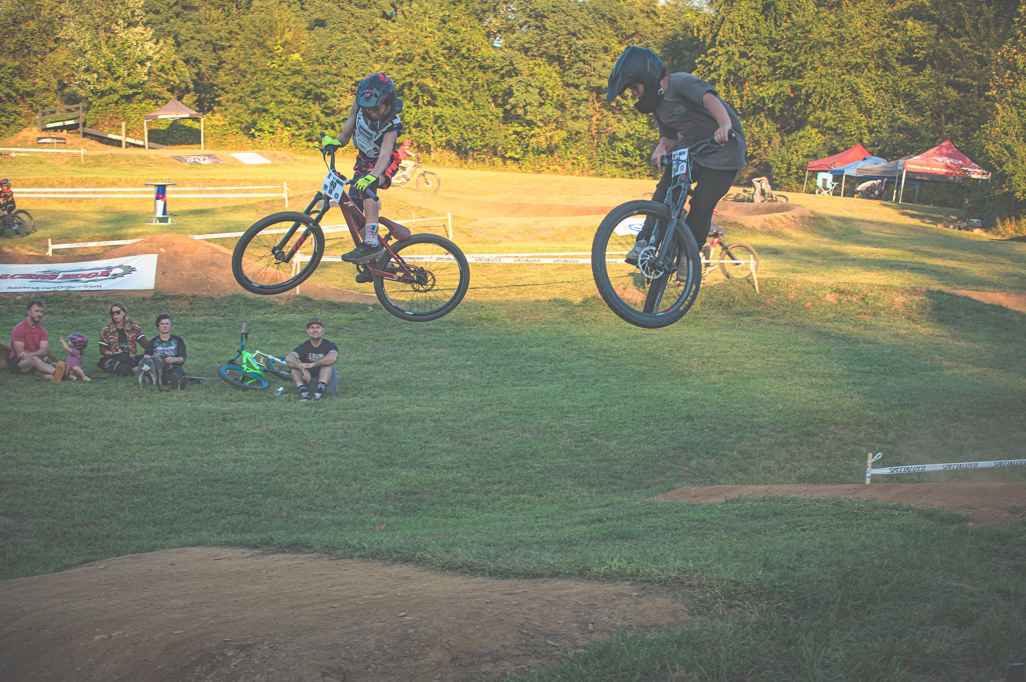 Two bikers performing jumps on a dirt track with spectators watching from a grassy area.