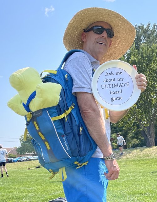 Man wearing sunglasses and a broad-brimmed hat, carrying a blue backpack and holding a frisbee that says "Ask me about my ULTIMATE board game" in a park setting.