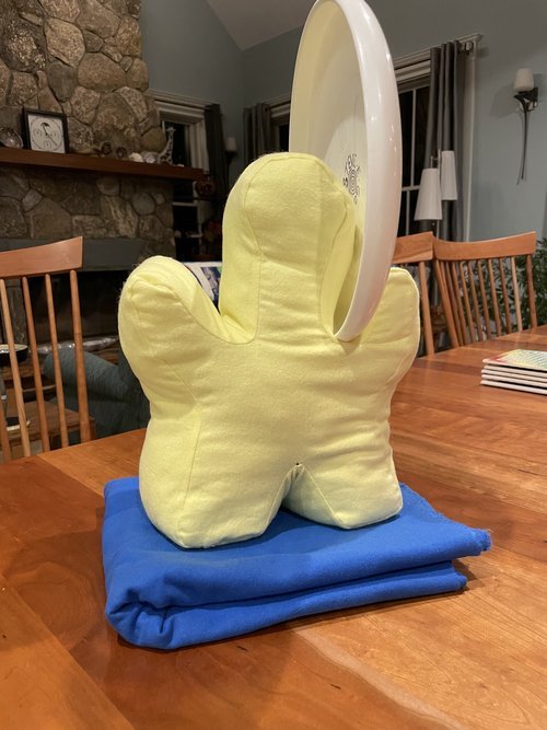 A plush yellow pillow shaped like a hand supporting a white frisbee, placed on a blue cloth on a wooden table. In the background, there are wooden chairs and a stone fireplace.