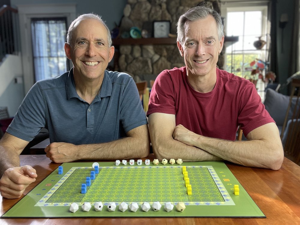 Two people smiling at a table with a board game in front of them, featuring green tiles and various game pieces and dice. The background shows a stone fireplace and a window.