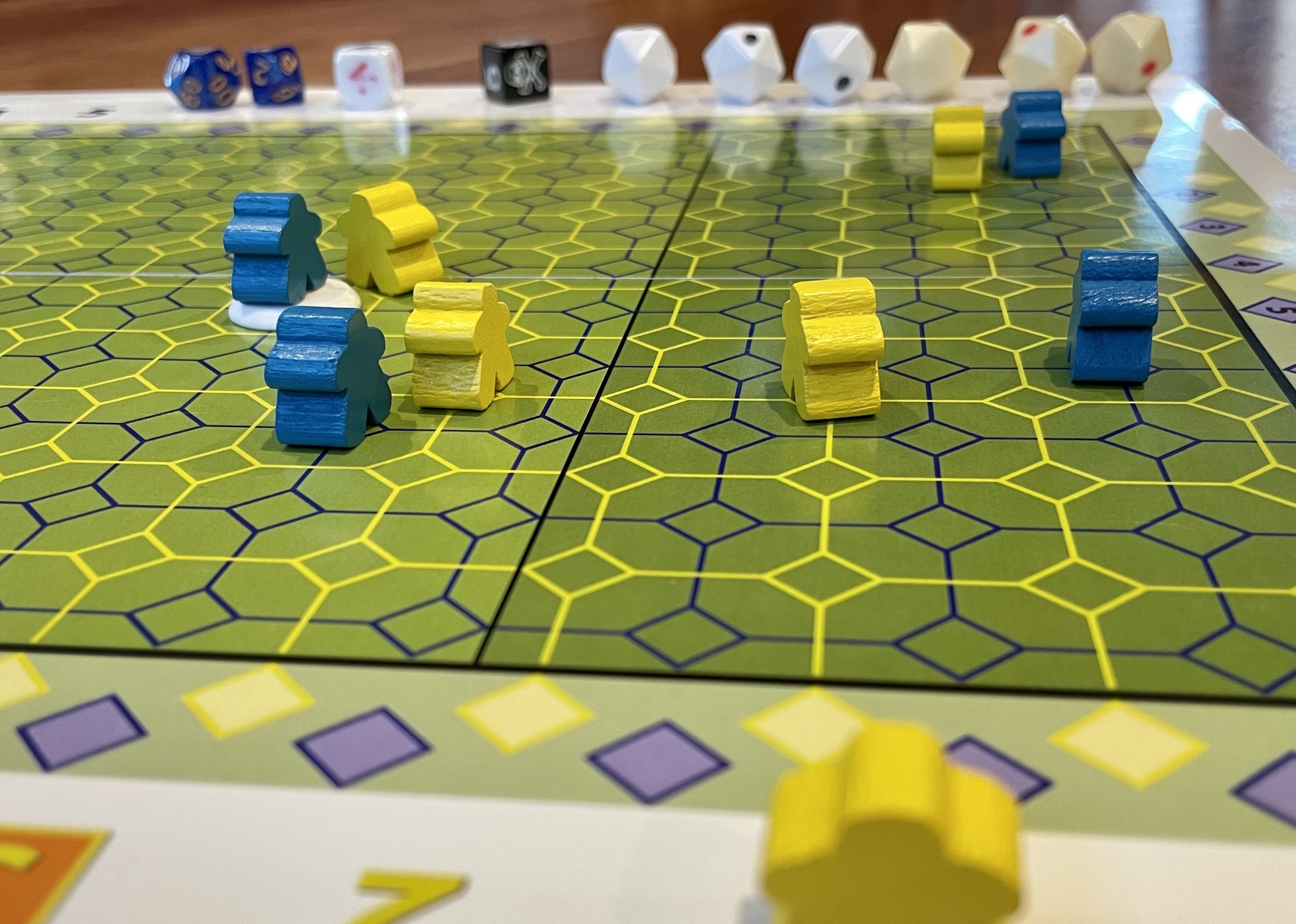 Board game with hexagonal grid and blue and yellow meeples on a green surface, accompanied by various dice in the background.