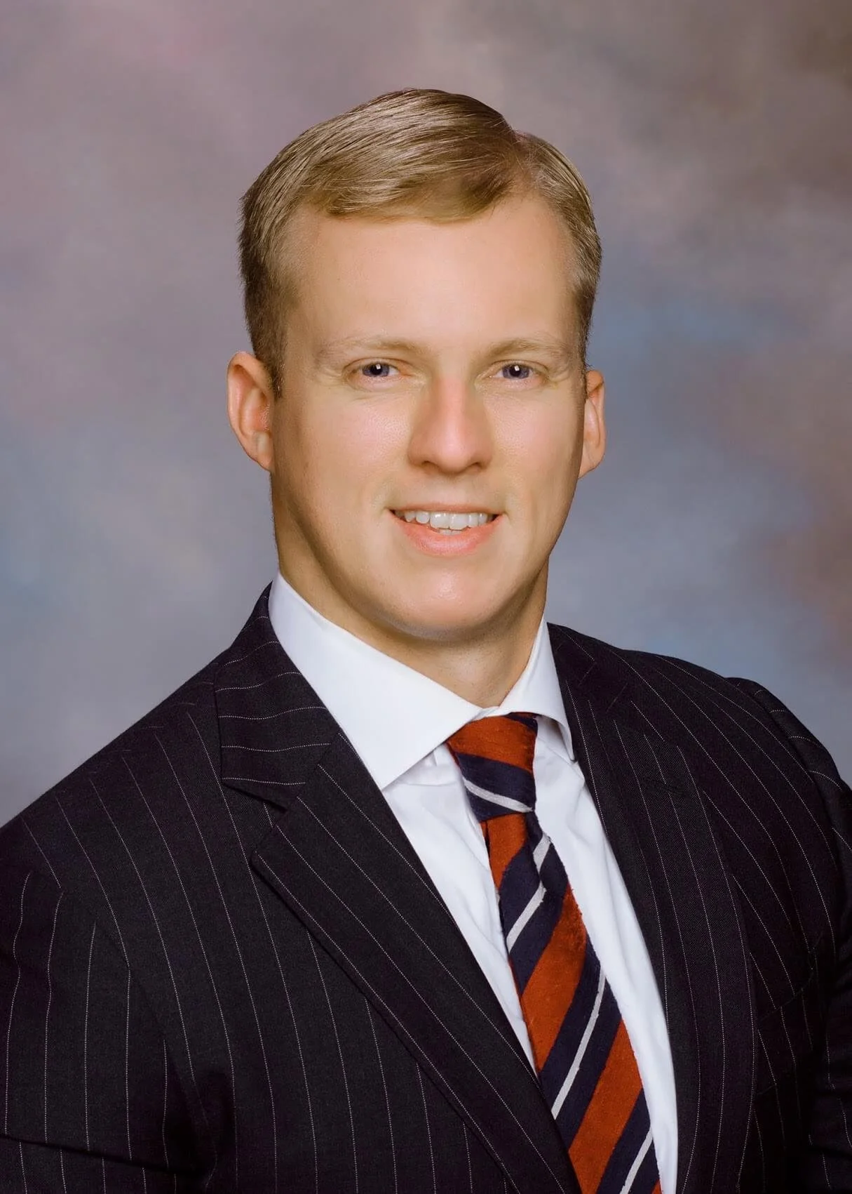 Headshot of Tom Gibson, smiling into the camera wearing a suit and red striped tie