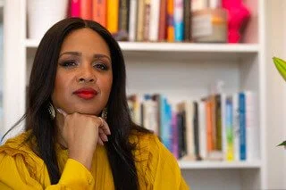 Headshot of Kimberly, looking confident wearing a mustard colored shirt in front of a bookshelf