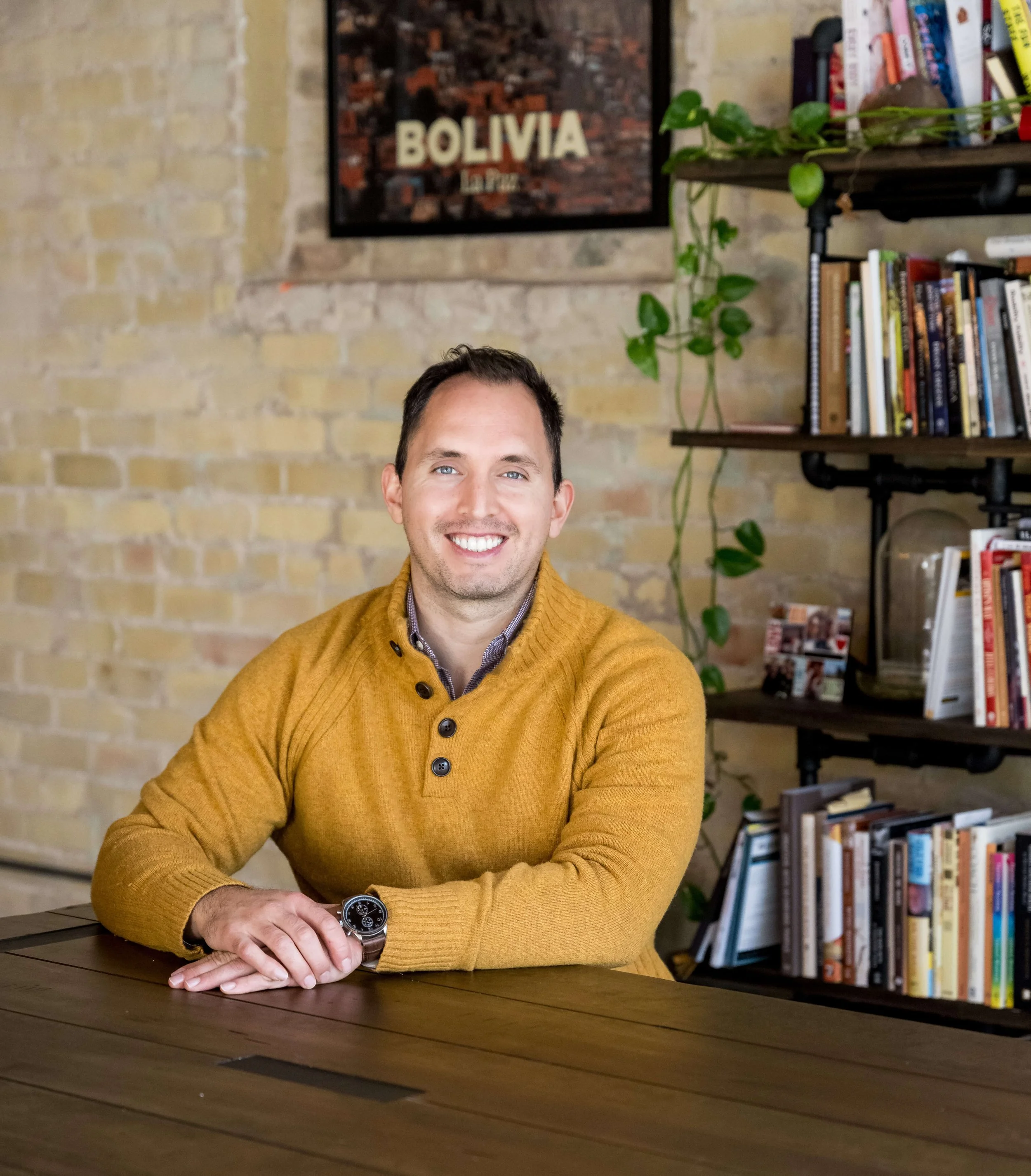 Derrick Deal smiles at the camera while sitting at a wooden table with books and plants on a shelf in the background