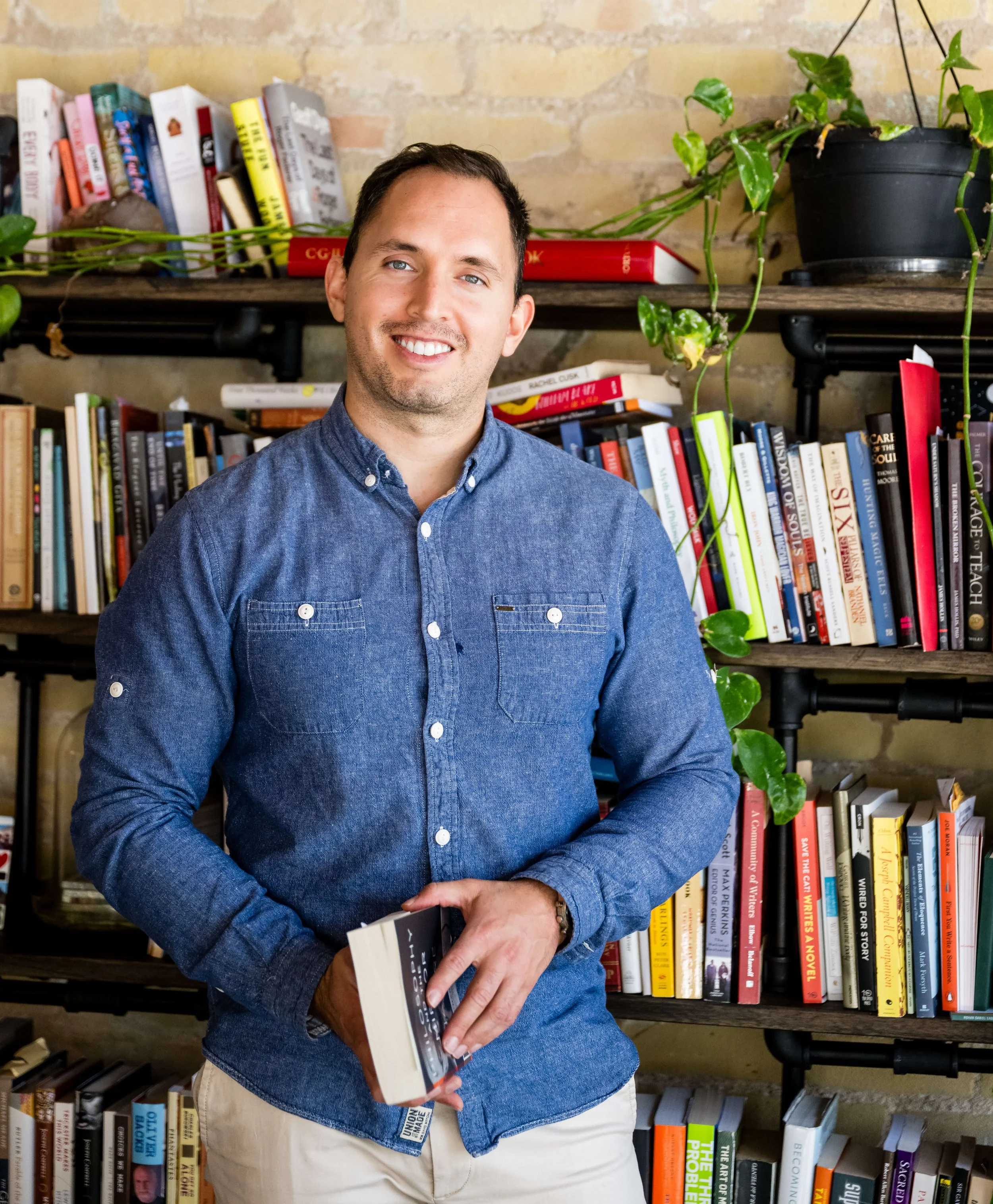 Derick Deal wears a blue denim shirt holding a book, standing in front of bookshelves filled with various books and a potted plant.