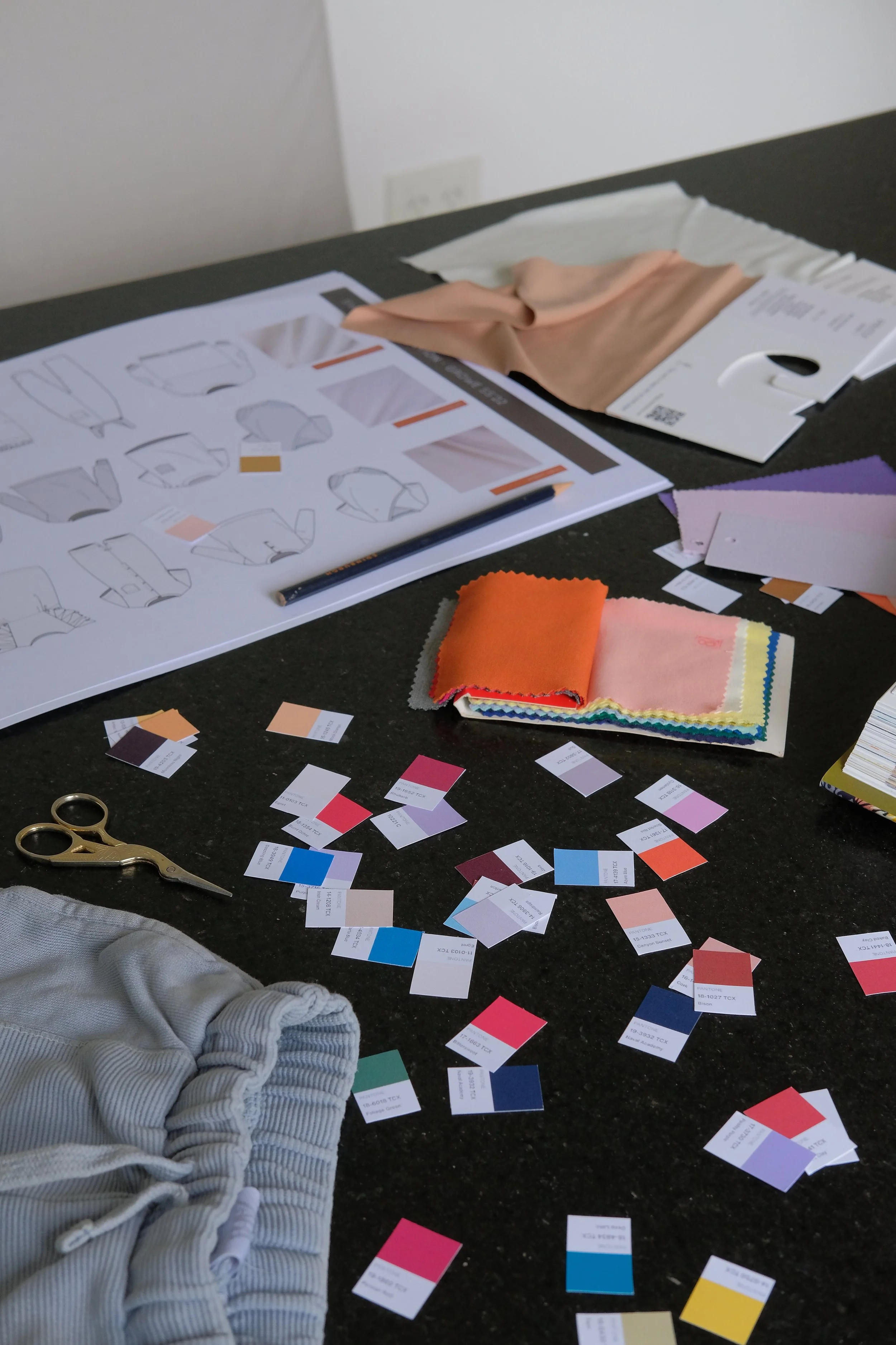 Color swatches, fabric samples, and design sketches for clothing on a black table in a workspace.