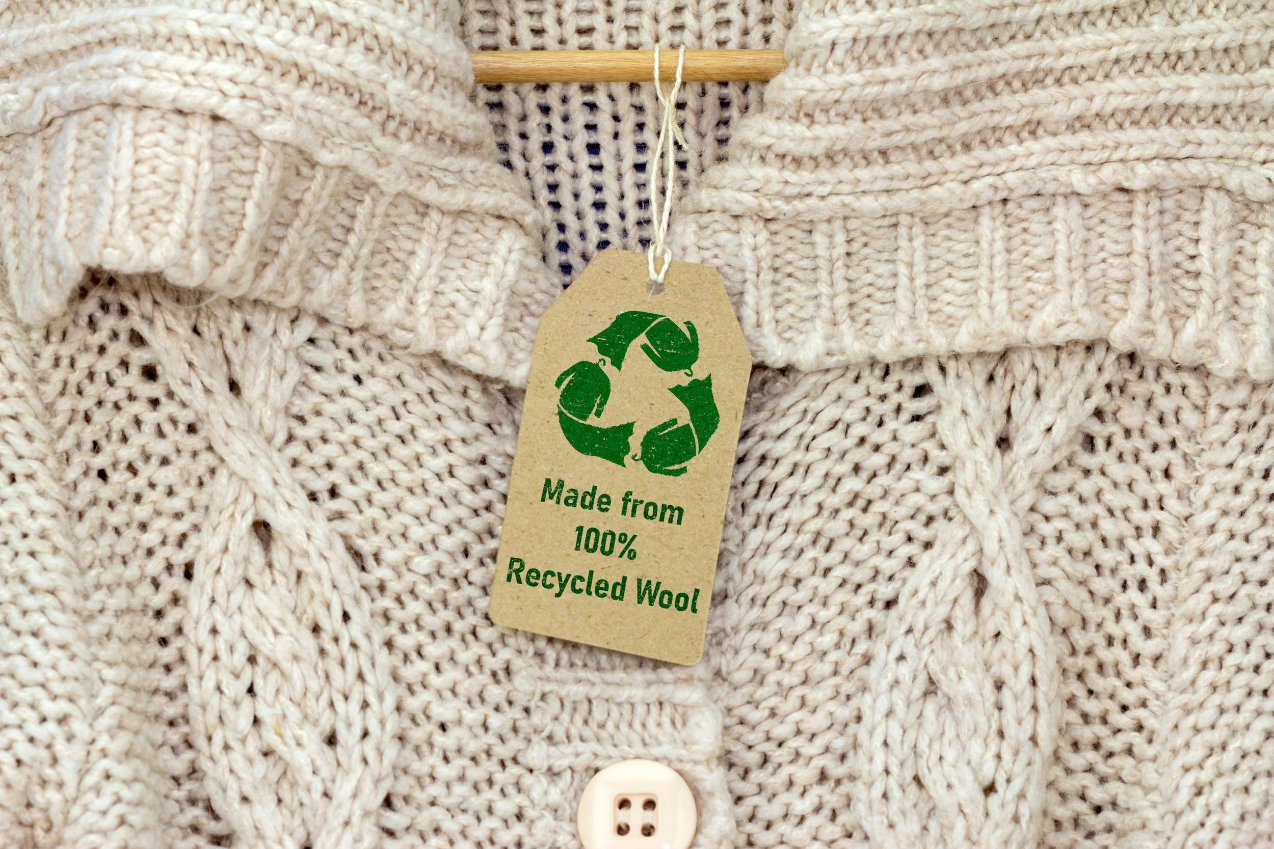Close-up of a knitted cream-colored sweater with a wooden hanger and a recycled wool tag attached with a string, showing a button at the bottom.