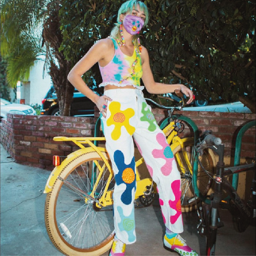 Person with teal hair wearing a colorful tie-dye crop top and white pants with large, colorful flower and swirl patterns, standing next to a yellow bicycle, wearing a purple face mask with a smiley face, in an outdoor setting with greenery and brick 