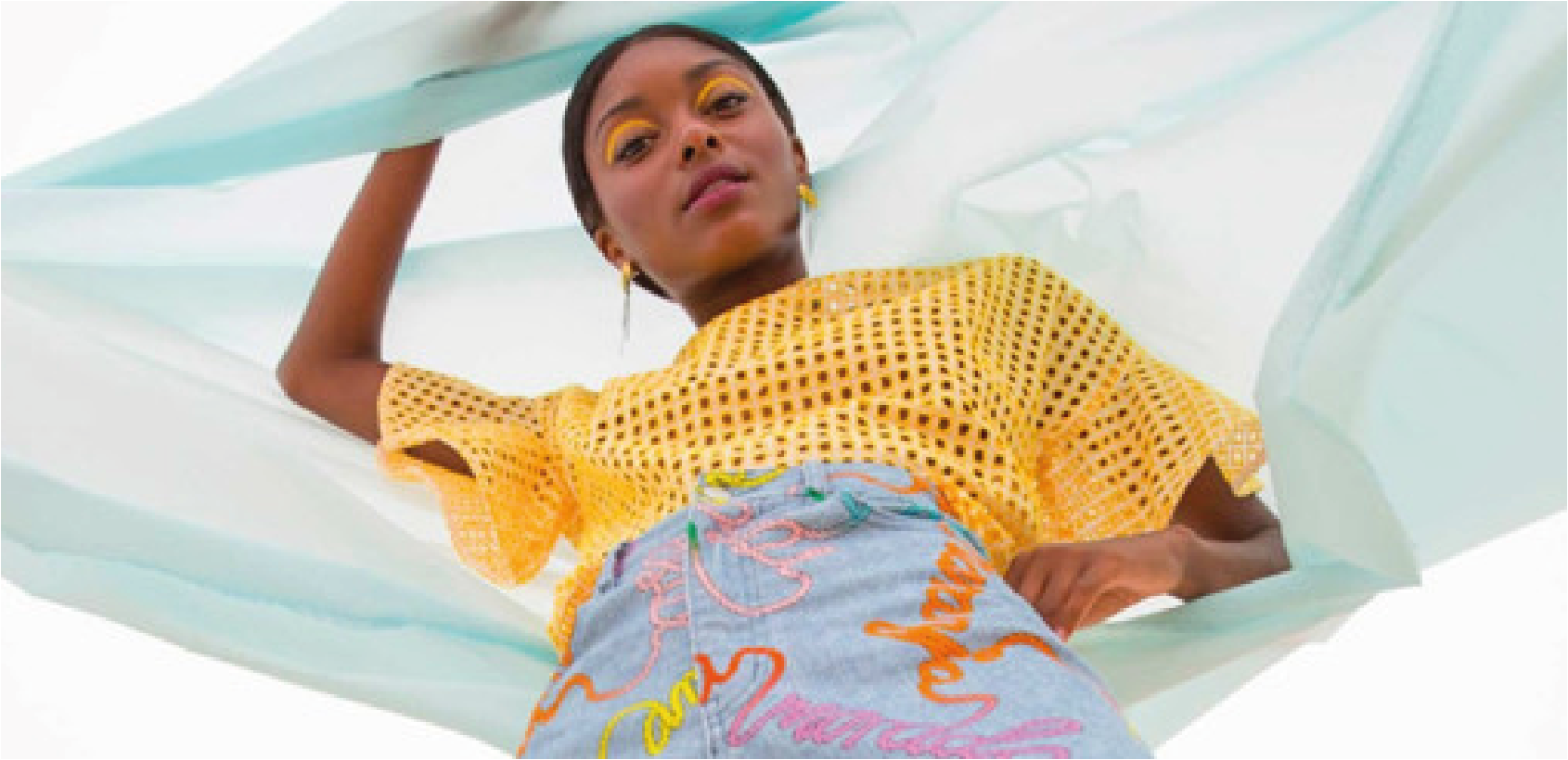 A woman looking down towards the camera, wearing bright yellow eye makeup, a yellow mesh top, and earrings, posed against a white background with a light blue sheer fabric around her.