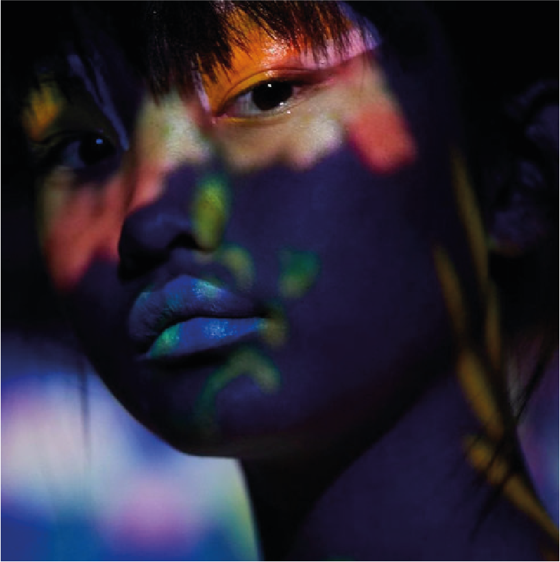 Close-up portrait of a woman with neon-colored paint on her face and lips, with colorful light reflections highlighting her features.