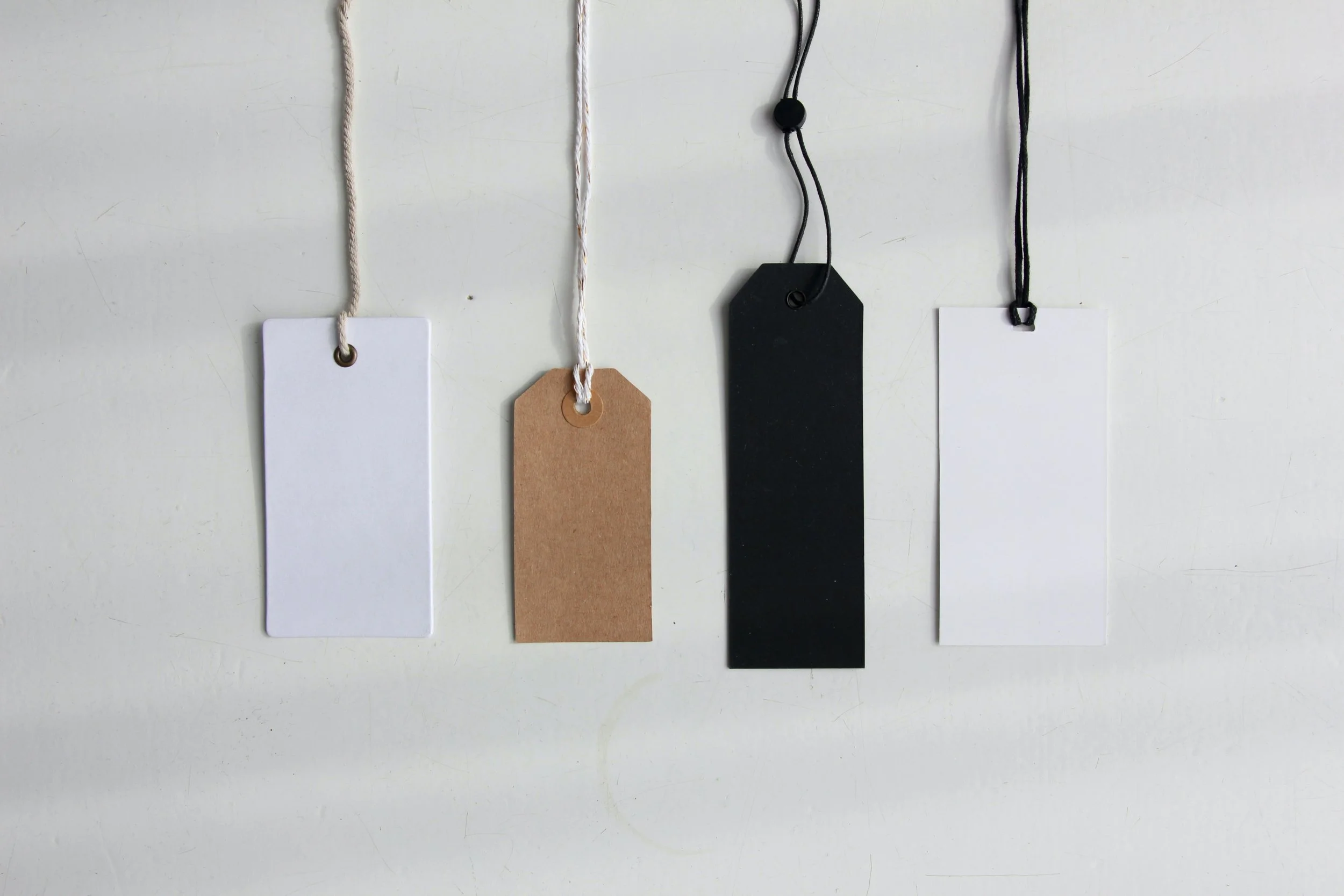 Four clothing tags hanging on a white background, each attached with a string of different colors and styles, including white, tan, black, and black with a white border.