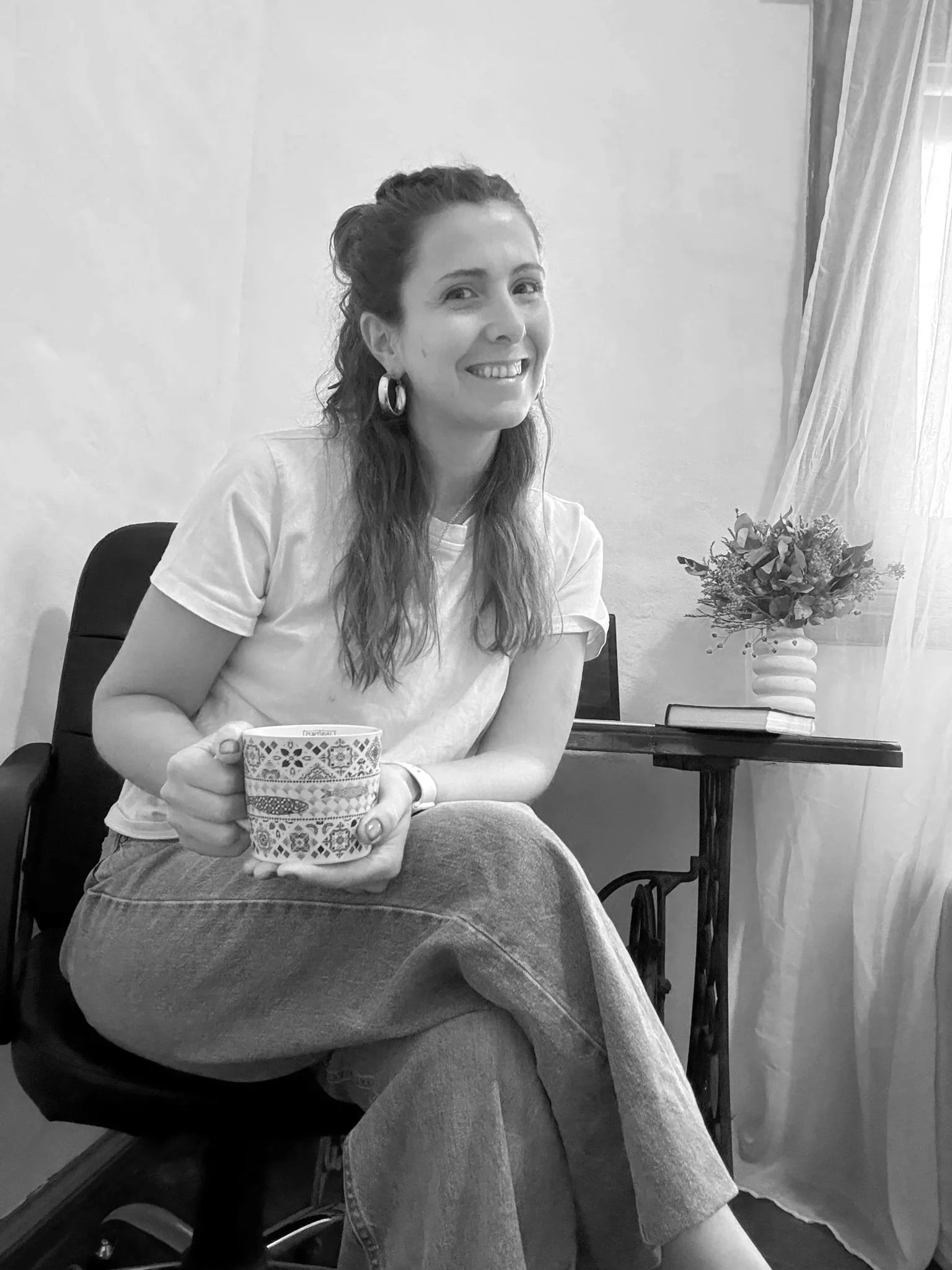 A woman sitting on a chair, smiling and holding a mug with a decorative pattern, in a room with a table, books, a vase with flowers, and a window with curtains.