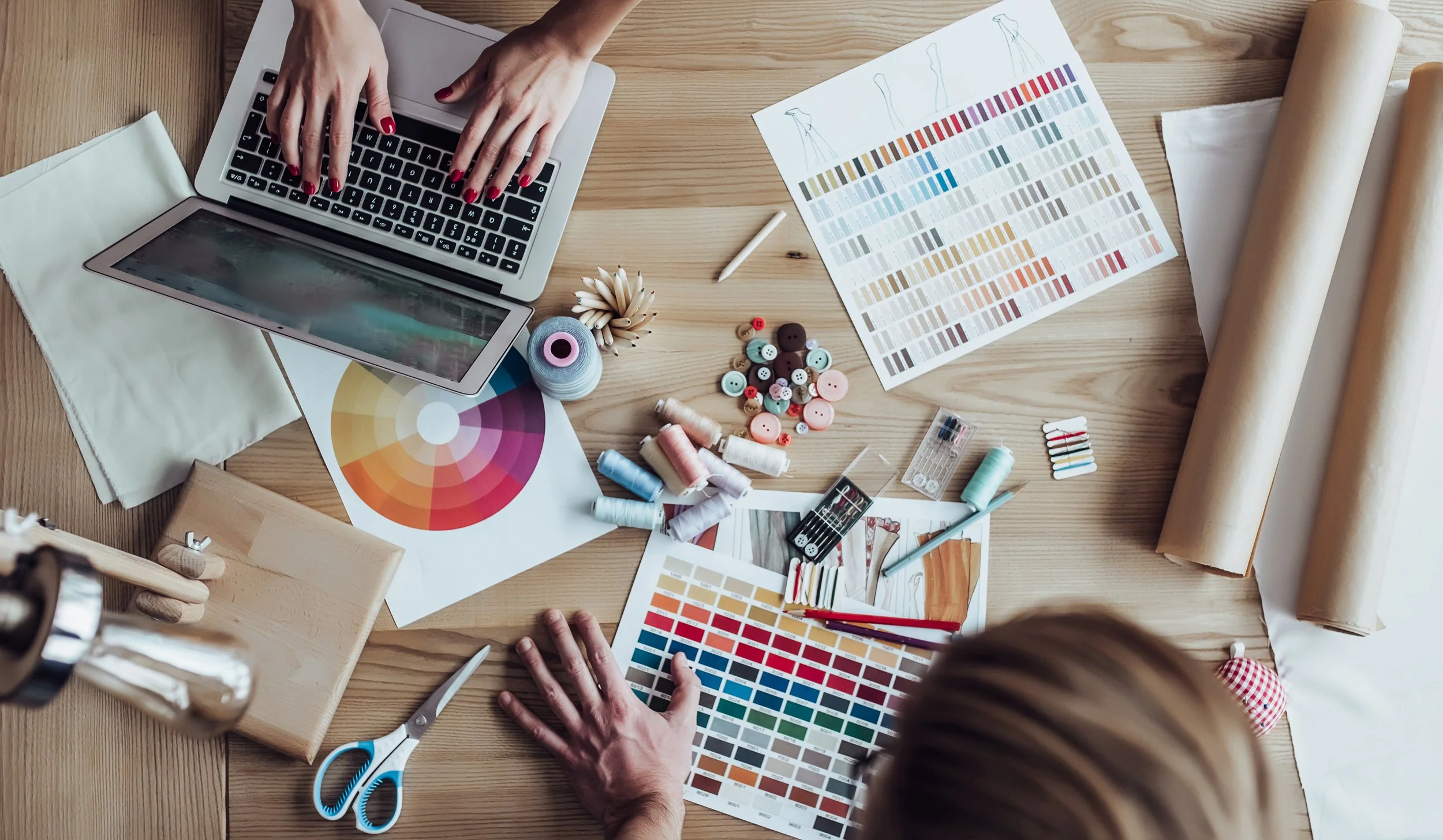 A workspace with a person's hands working on a laptop, surrounded by color swatches, fabric rolls, buttons, thread spools, scissors, and design sketches, indicating a fashion or textile design project.