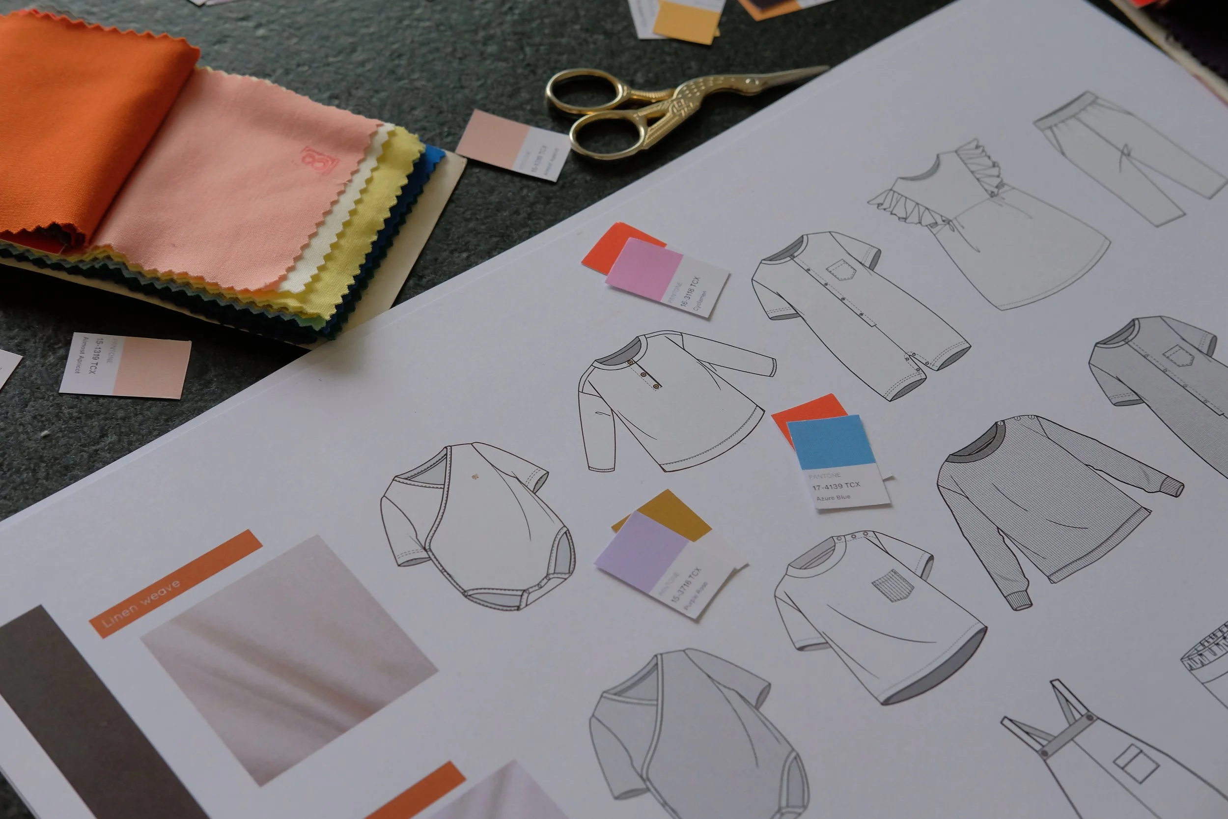 Fashion design sketches of a shirt with detailing, color swatches, fabric samples, and scissors on a table.
