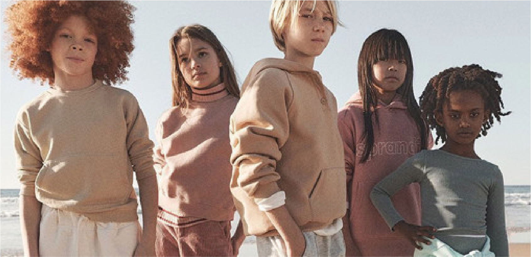 Five children standing on the beach with ocean waves in the background, wearing casual clothing.