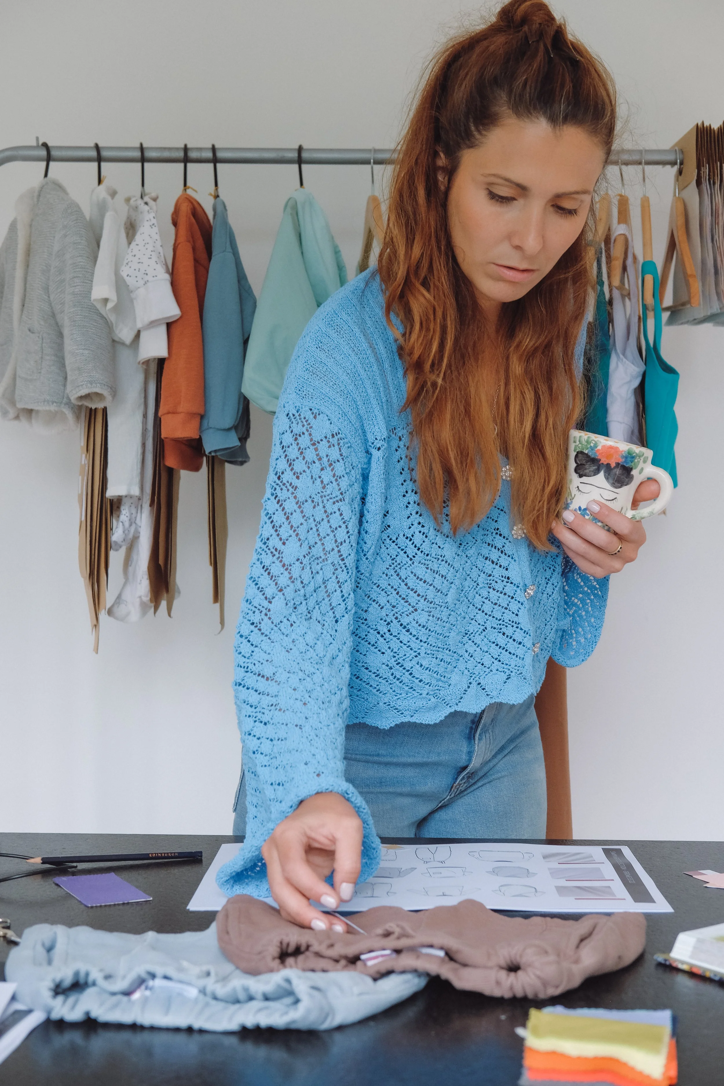 Woman with long red hair wearing a blue crochet sweater, holding a coffee mug with sunglasses and floral design. She is looking down at a table with fabric, paper, and sewing supplies. Behind her, a clothing rack with various shirts and garments hanging.