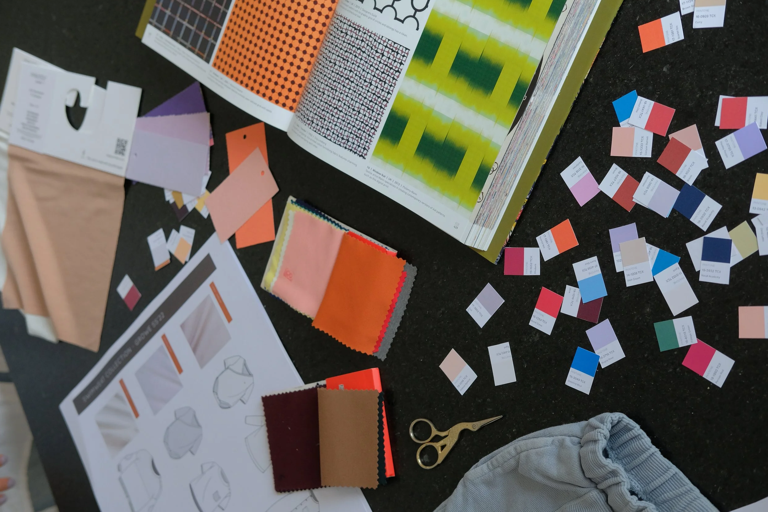 Color swatches, fabric samples, printed color charts, scissors, and pattern sketches on a black surface for textile or fashion design.