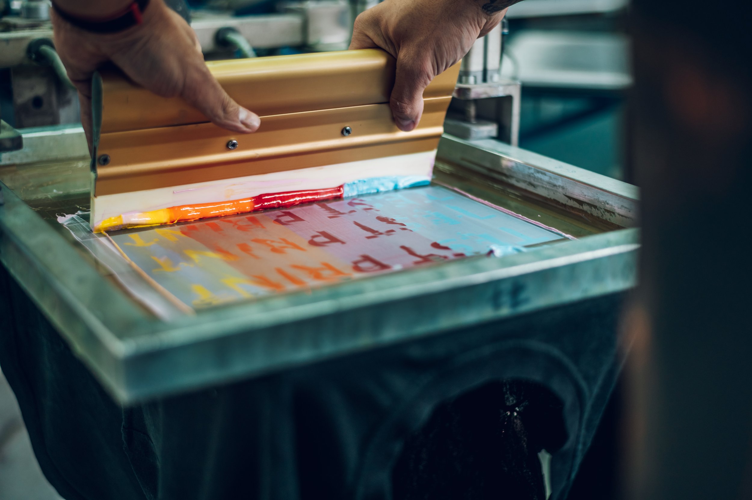 Close-up of hands operating a printing or screen printing machine with colorful ink on a flat surface.
