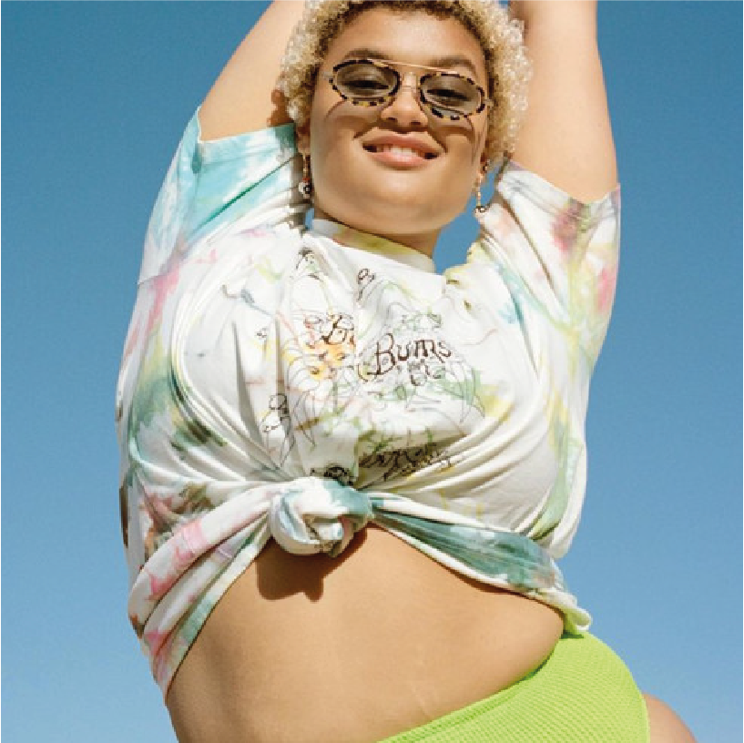 Woman with curly blonde hair wearing sunglasses, a tie-dye shirt tied at the side, and bright lime green shorts, standing against a clear blue sky.