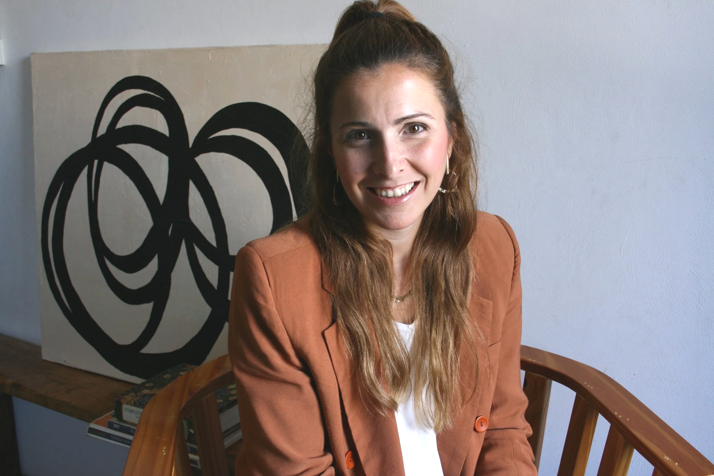 A woman with long, light brown hair tied in a high ponytail, smiling, wearing a brown blazer over a white top, sitting on a wooden chair in front of a white wall with a black abstract painting of a rose behind her.