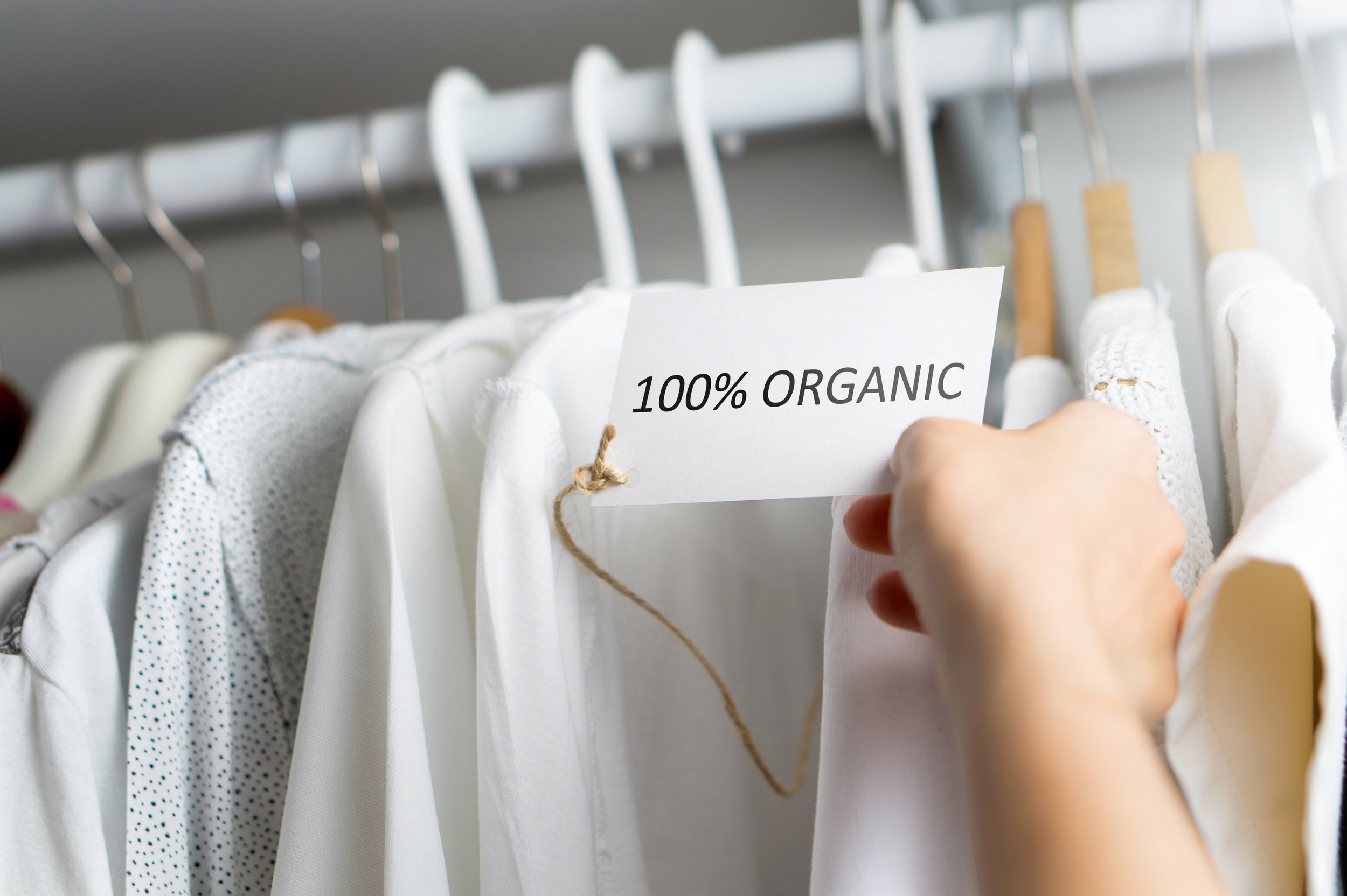 Person holding a white card with black text reading '100% Organic' in front of clothing items hanging on a rail, including white and patterned garments.