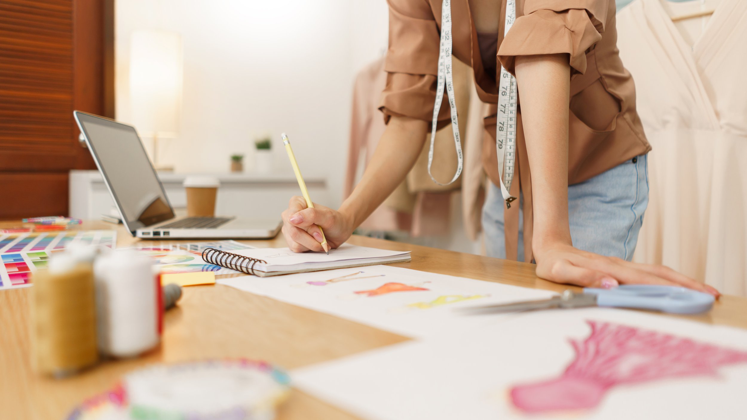 Person leaning over a table, writing in a notebook with a pencil, surrounded by art supplies and color samples.
