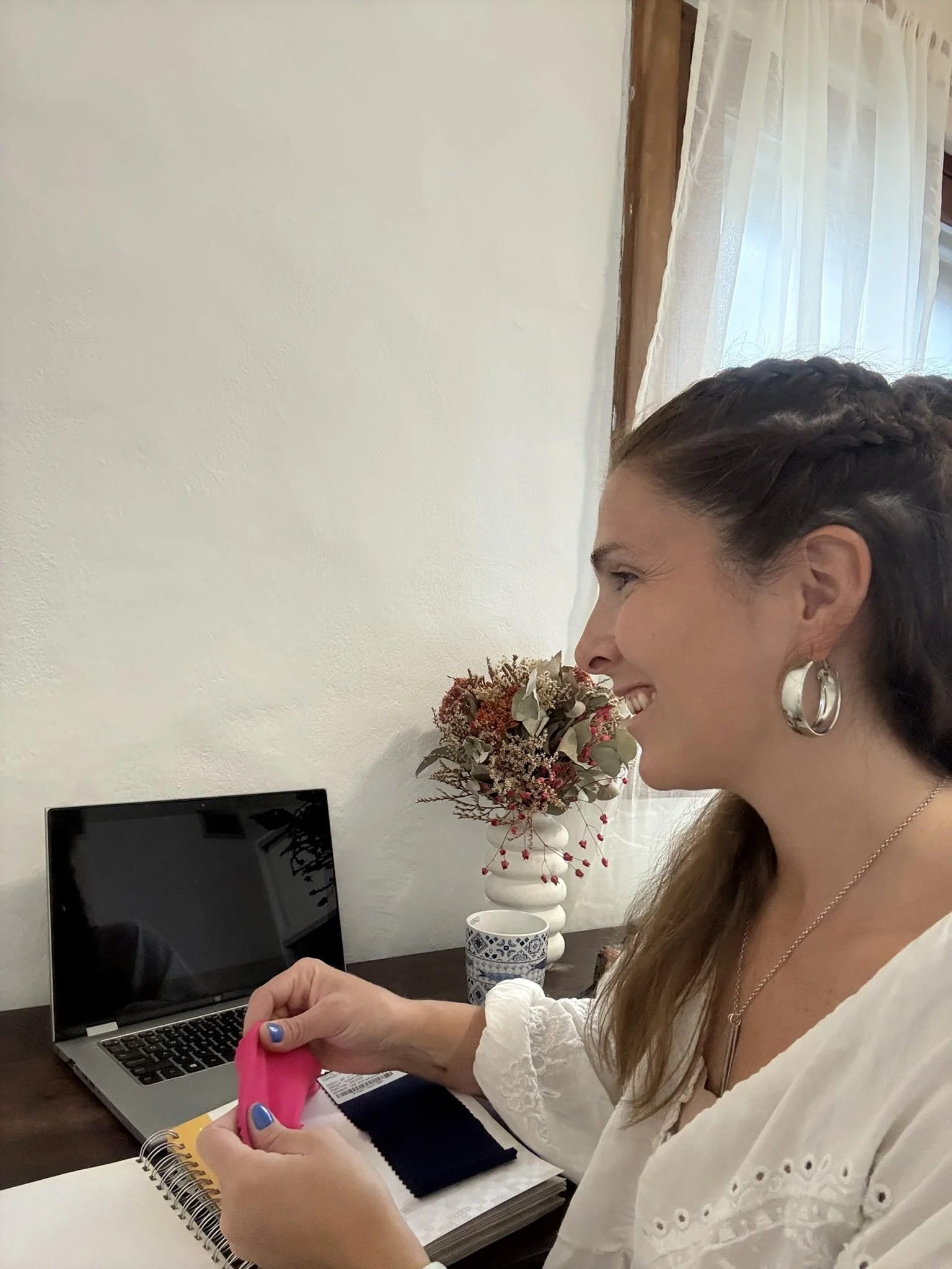 A woman smiling while looking at her phone, sitting at a desk with an open laptop, a notebook, and a cup in front of her. There is a bouquet of flowers on the desk and a window with sheer curtains in the background.