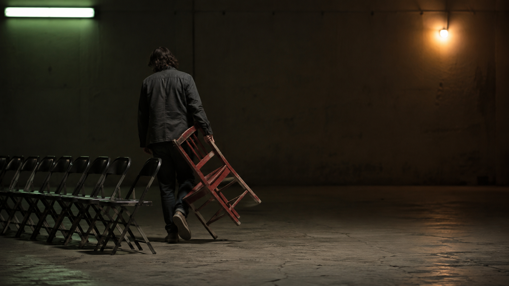 A figure carries a red chair away from a rigid row, leaving a gap under green and amber practical light.