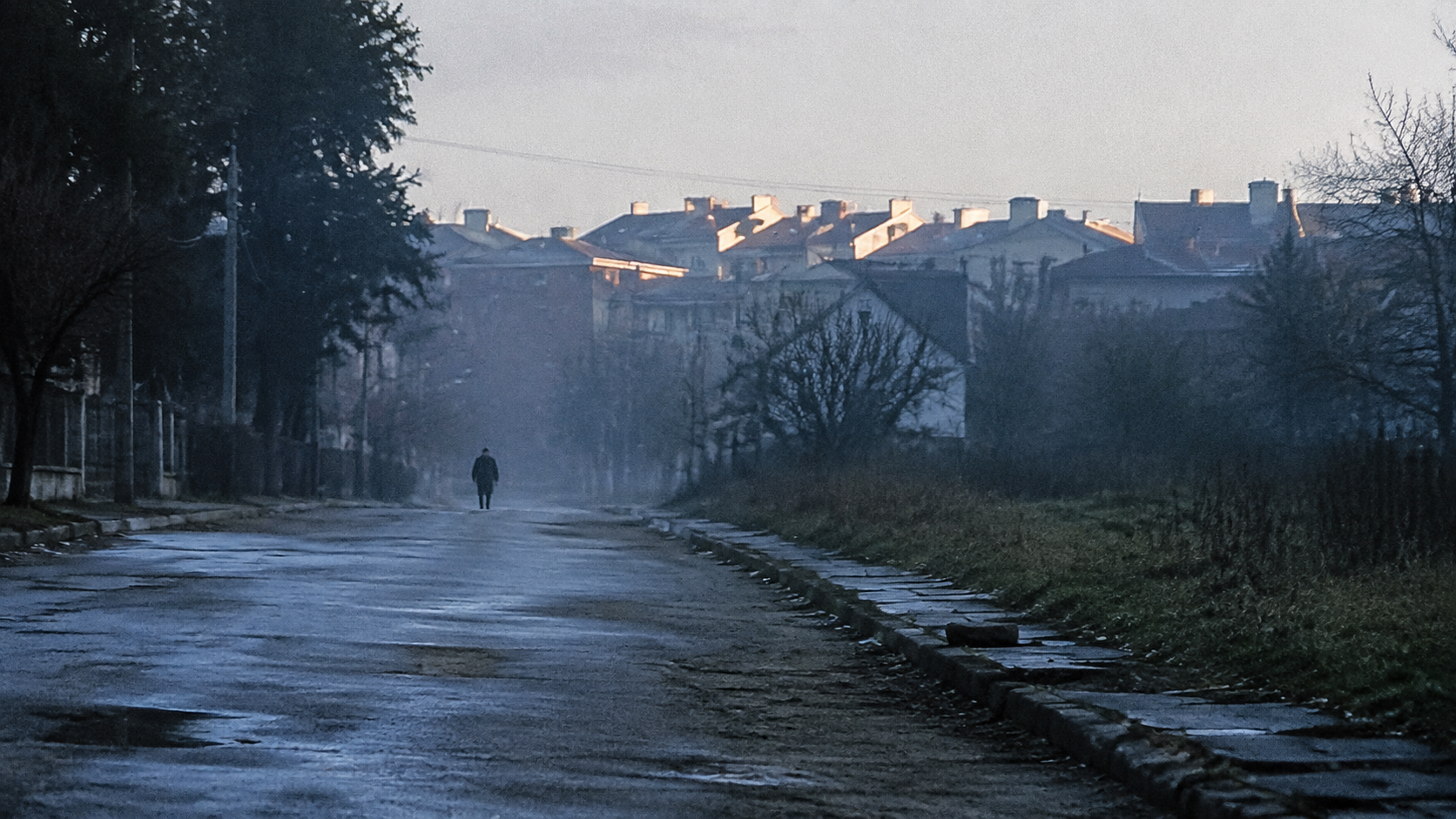 A tiny figure approaches along an empty dawn street beneath faint haze and pale light on distant rooftops.