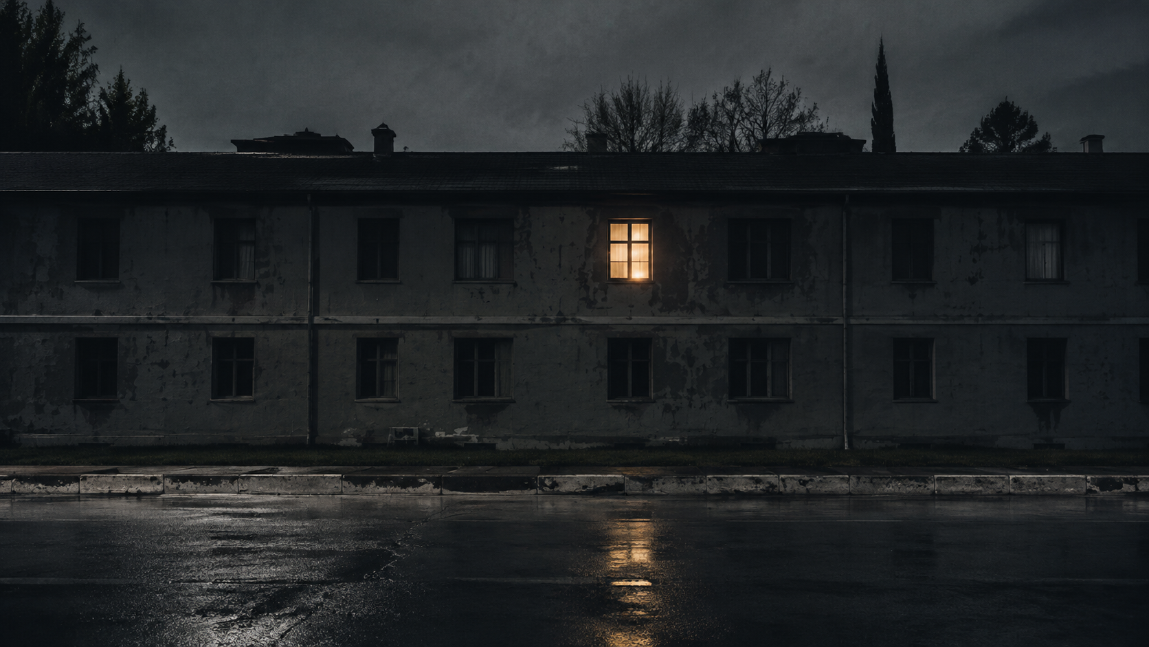 A dark apartment building across a wet street has one warm window lit on the upper floor.