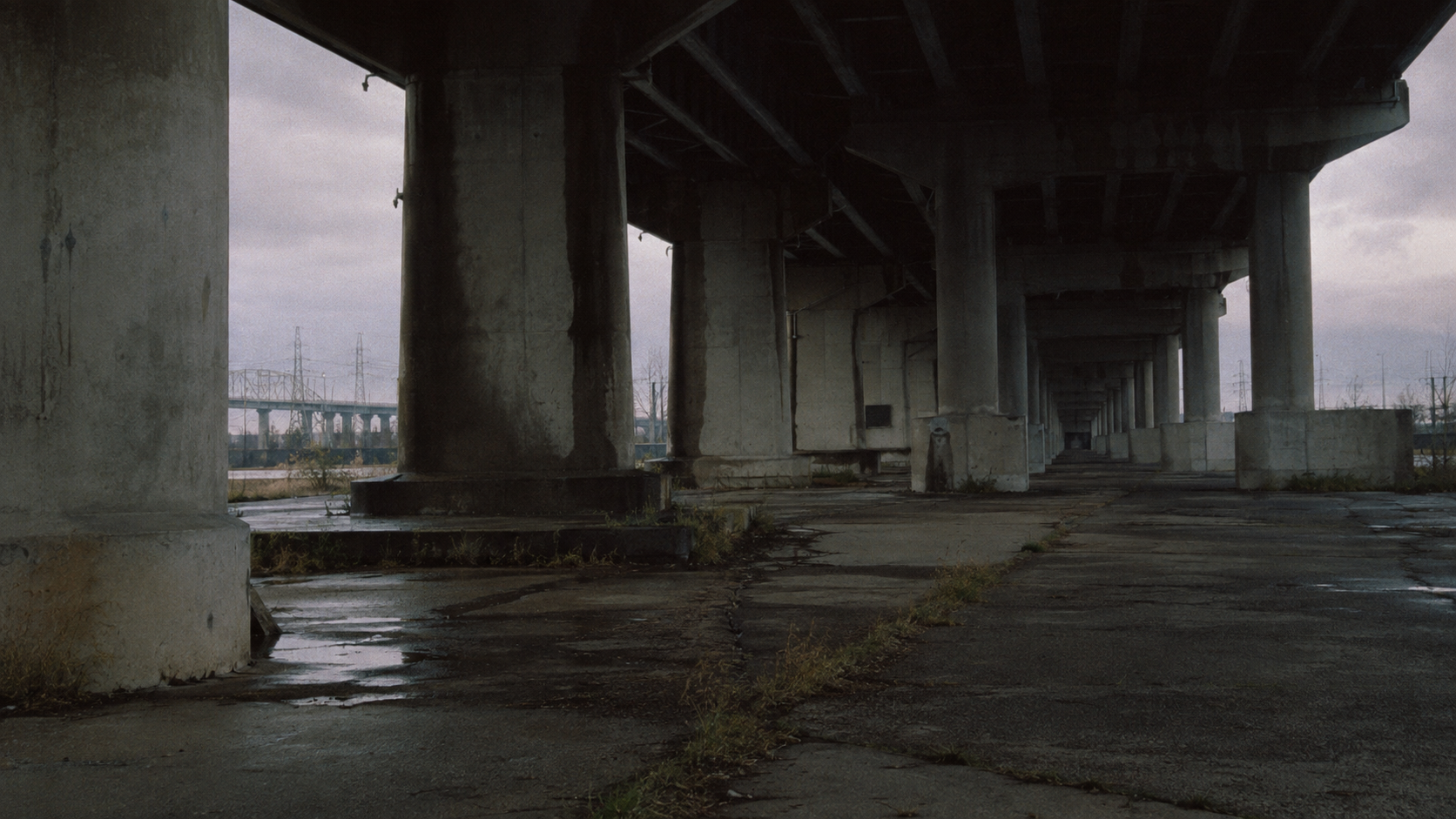 Weather-stained concrete supports recede beneath an empty highway overpass in flat dusk light.