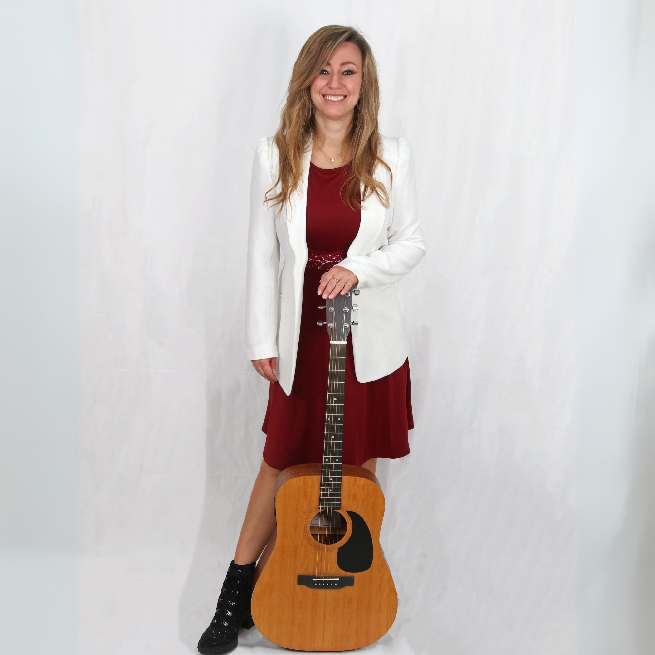 Eine Frau mit langem, gewelltem Haar steht vor einem weißen Hintergrund, trägt eine weiße Jacke über einem roten Kleid und lacht. Sie hält eine Akustikgitarre, die auf dem Boden steht, mit einer Hand. Sie trägt schwarze Stiefel.